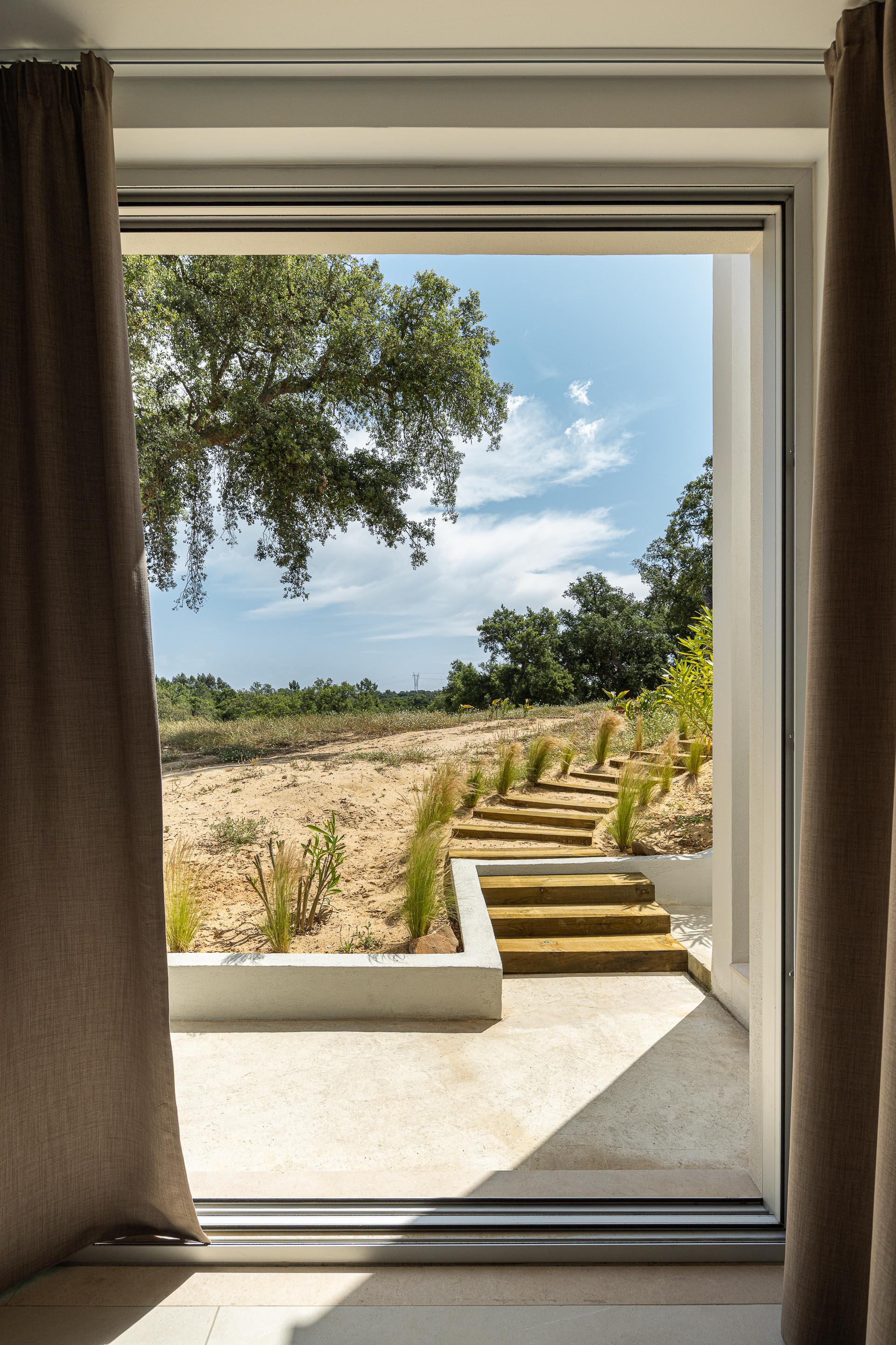 Vão envidraçado com cortinas, abrindo para degraus em madeira natural e um jardim árido com vegetação — Casa M Ariane fotografia Ivo Tavares