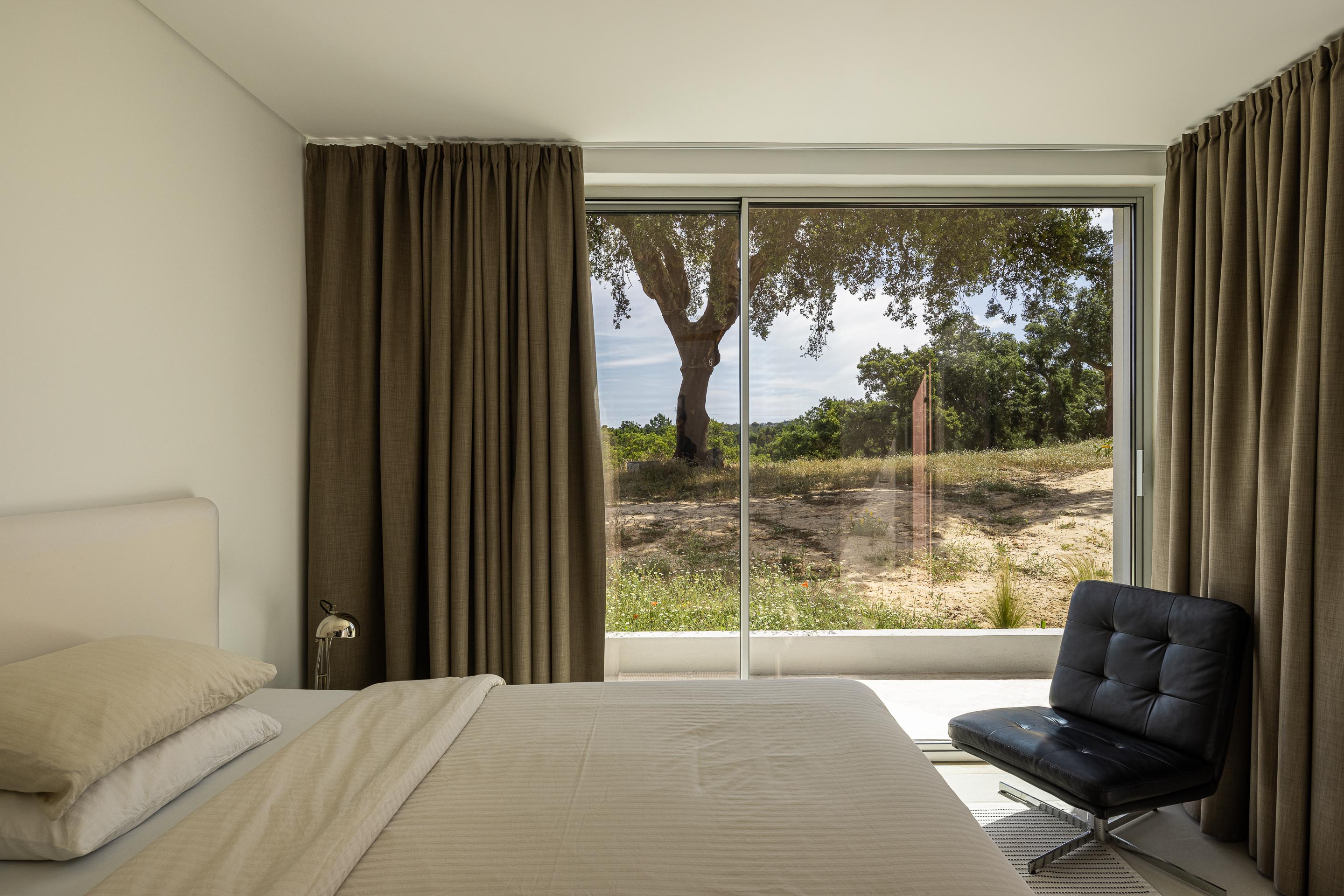 Quarto minimalista com pavimento claro, grande vão envidraçado e vista para a paisagem. Poltrona de design em pele negra — Casa M Ariane fotografia Ivo Tavares