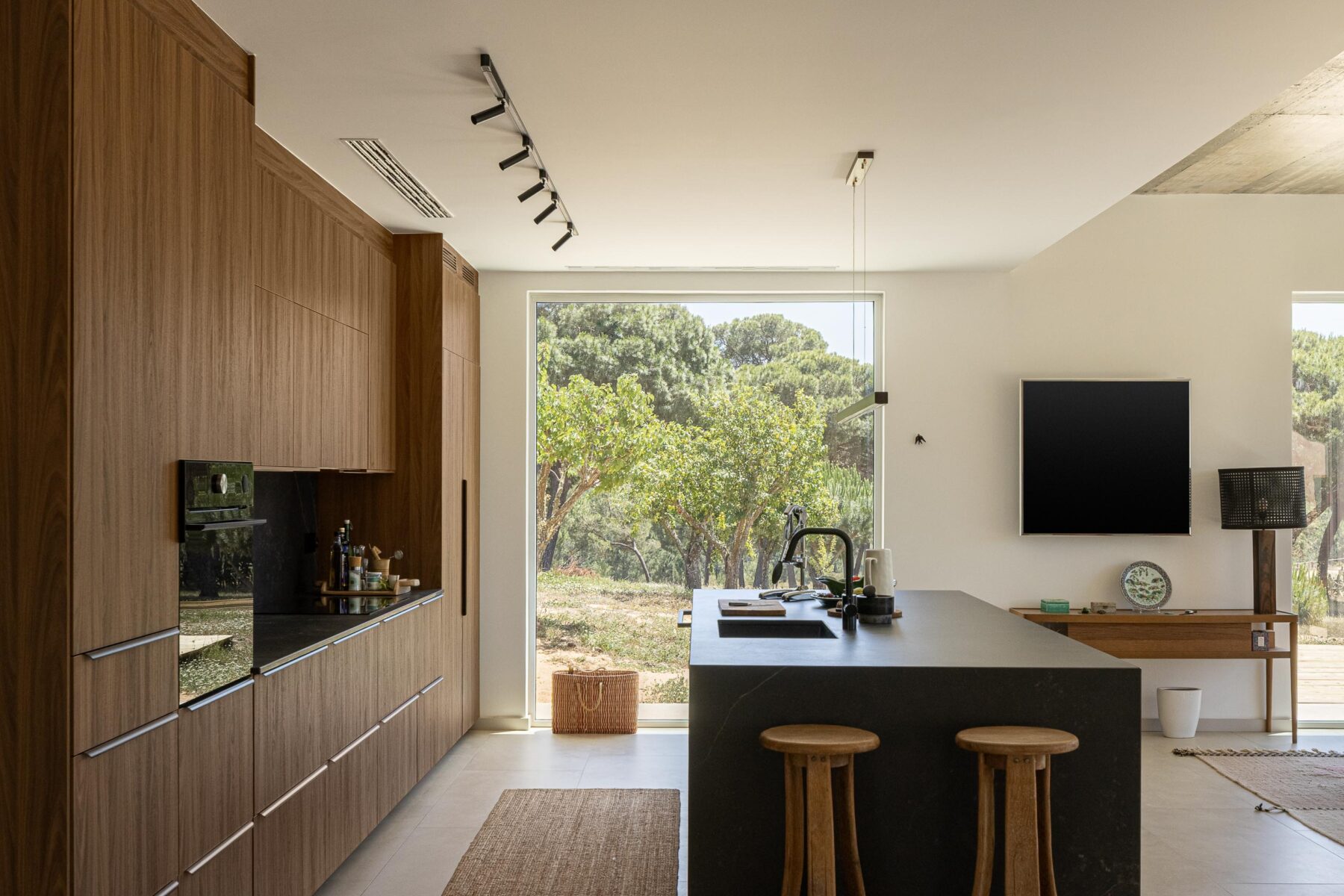 Cozinha com armários em madeira escura, ilha em pedra negra e pavimento em cerâmica clara, com vista para a natureza — Casa M Ariane fotografia Ivo Tavares