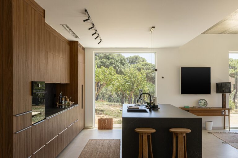 Cozinha com armários em madeira escura, ilha em pedra negra e pavimento em cerâmica clara, com vista para a natureza — Casa M Ariane fotografia Ivo Tavares