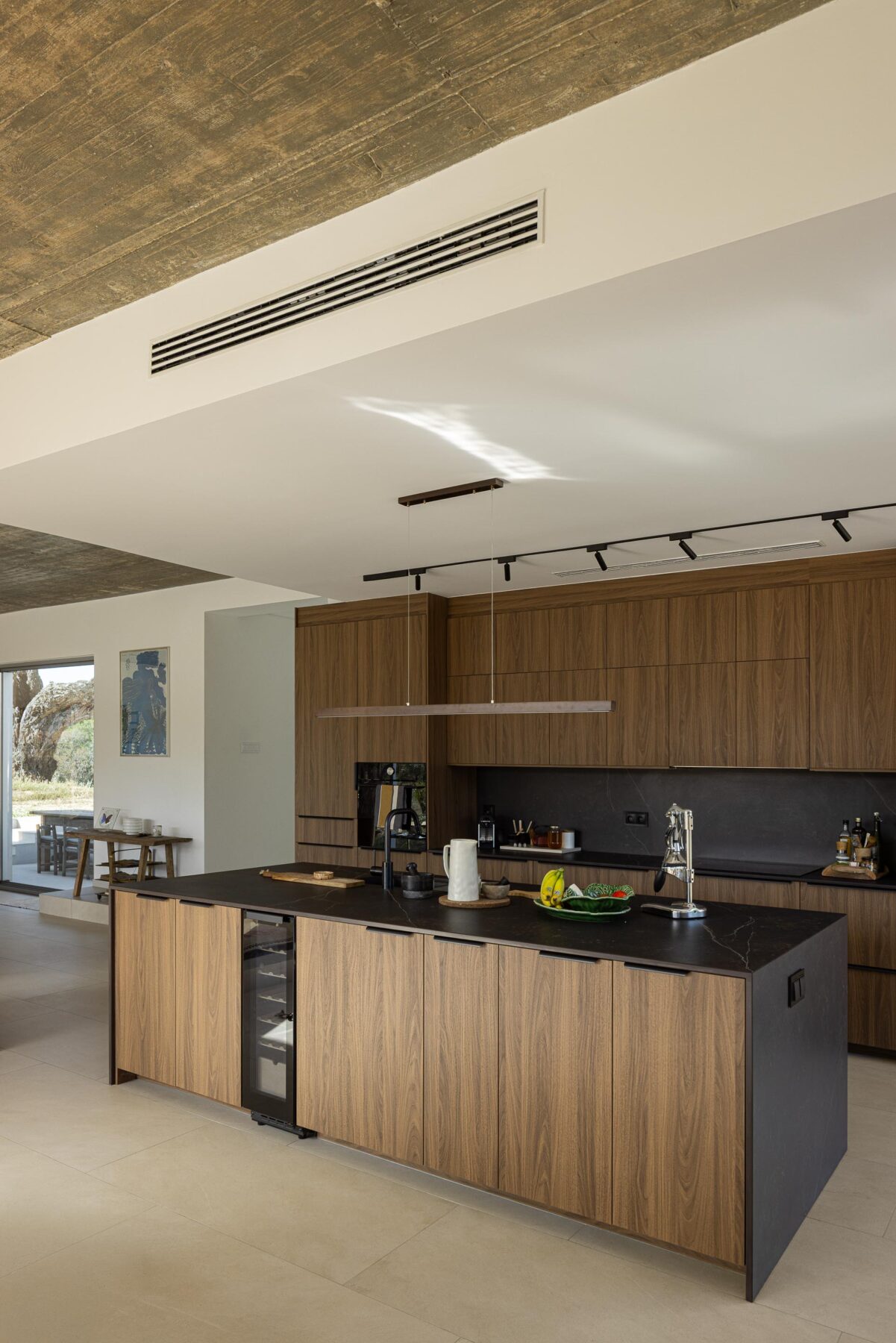 Cozinha com armários em madeira escura, ilha em pedra negra e pavimento claro, sob cobertura em betão aparente — Casa M Ariane fotografia Ivo Tavares
