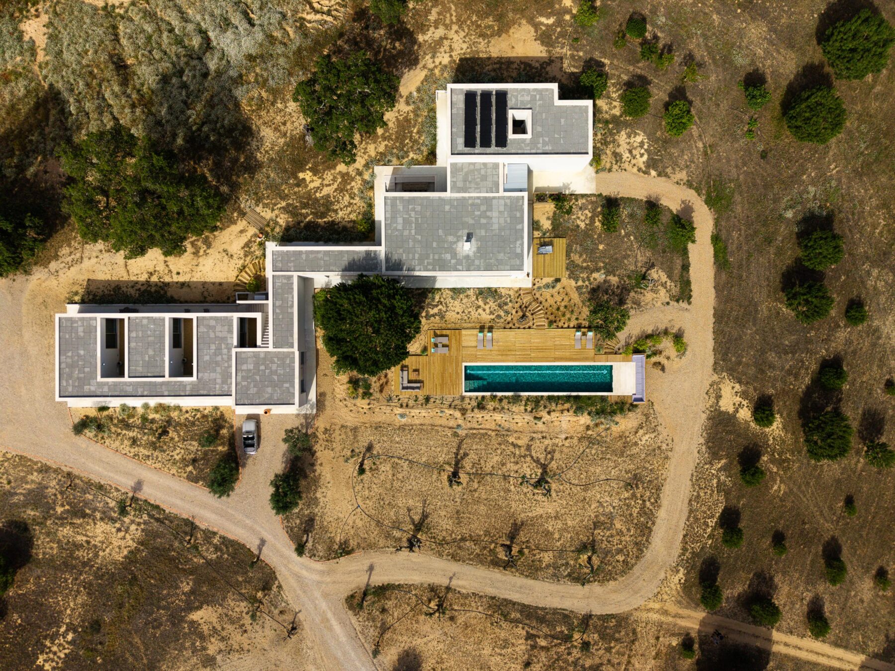 Vista aérea da moradia em planta L com coberturas planas, piscina e deck em madeira, integrada na paisagem árida — Casa M Ariane fotografia Ivo Tavares