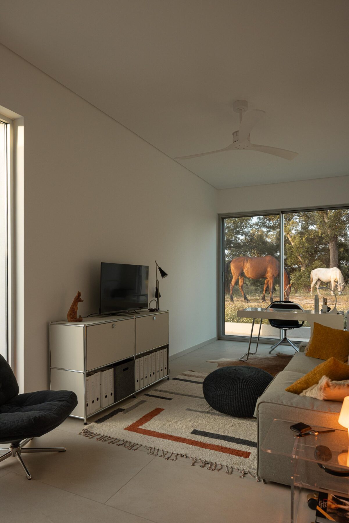 Sala de estar com pavimento claro e paredes brancas, com grande vão envidraçado e vista para cavalos no exterior — Casa M Ariane fotografia Ivo Tavares