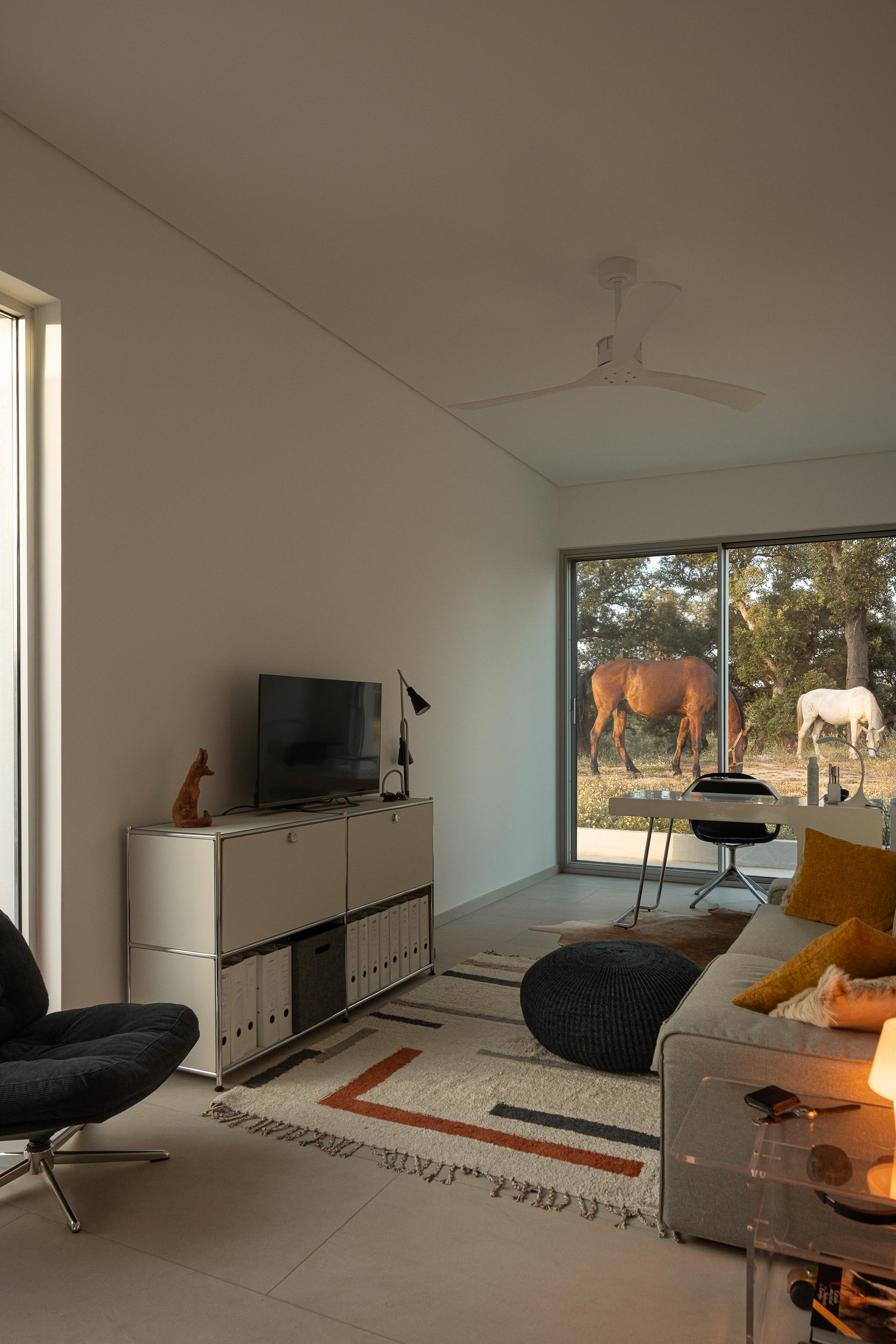Sala de estar com pavimento claro e paredes brancas, com grande vão envidraçado e vista para cavalos no exterior — Casa M Ariane fotografia Ivo Tavares