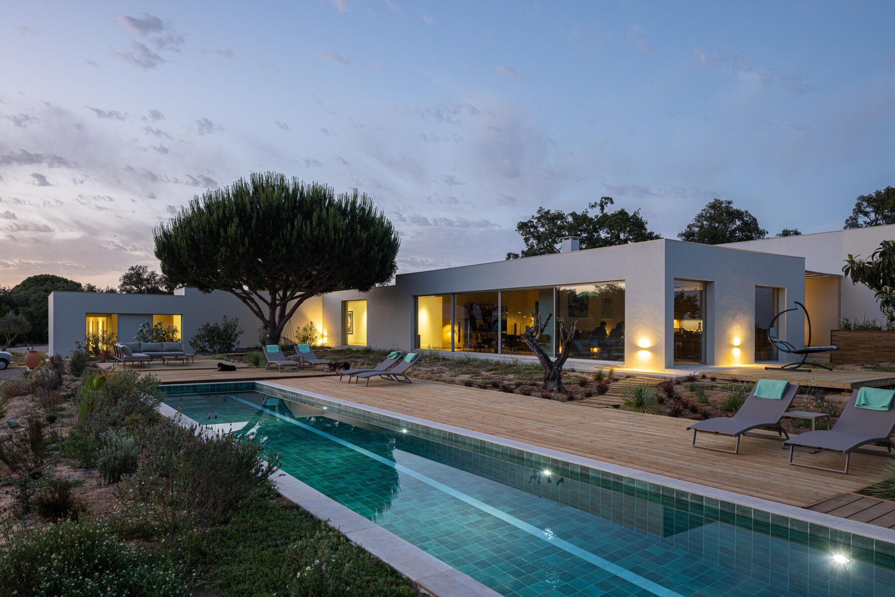 Exterior da moradia com fachada branca e grandes vãos iluminados, deck em madeira e piscina à noite — Casa M Ariane fotografia Ivo Tavares