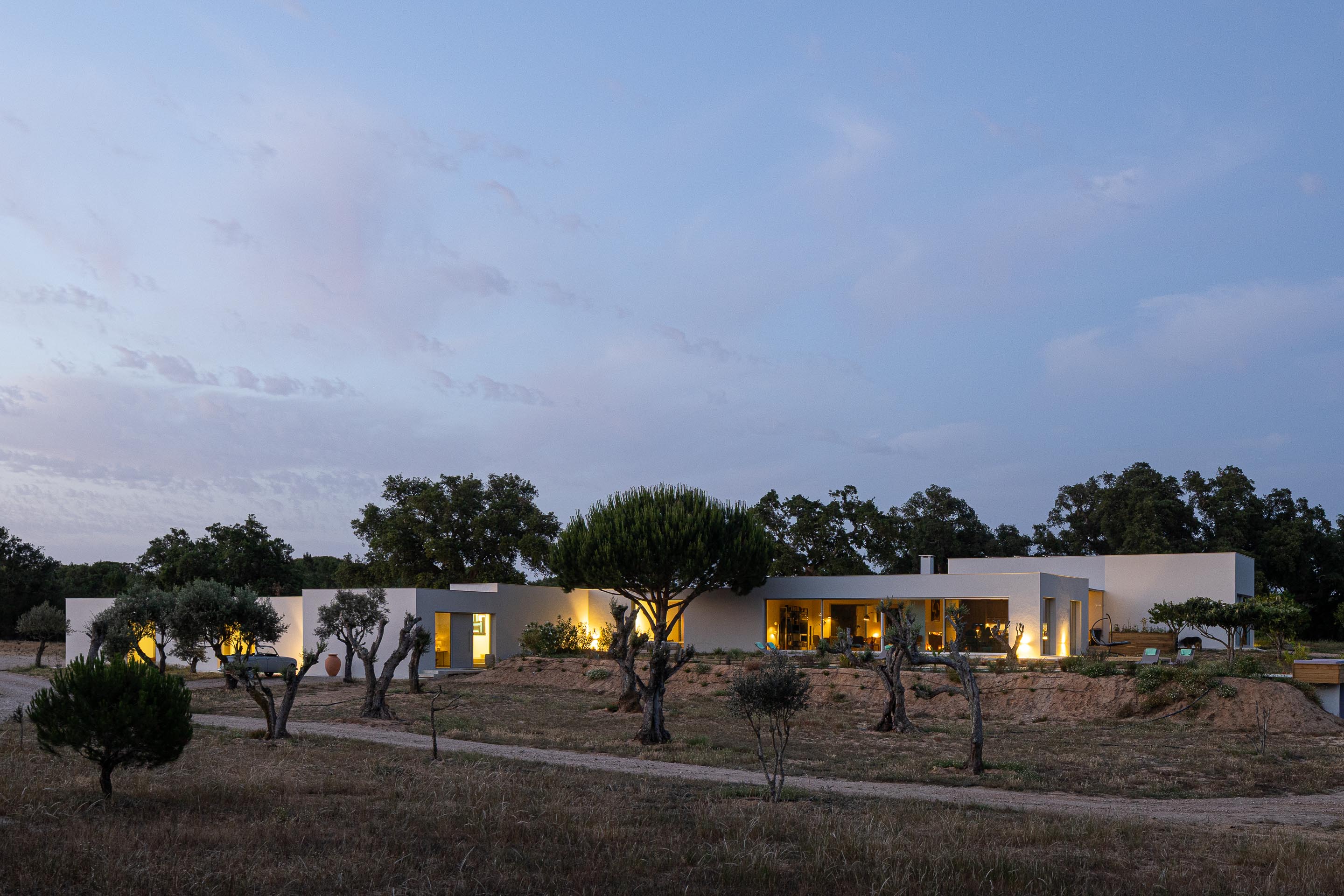 Vista geral da moradia branca com vãos iluminados, integrada na paisagem alentejana ao anoitecer — Casa M Ariane fotografia Ivo Tavares