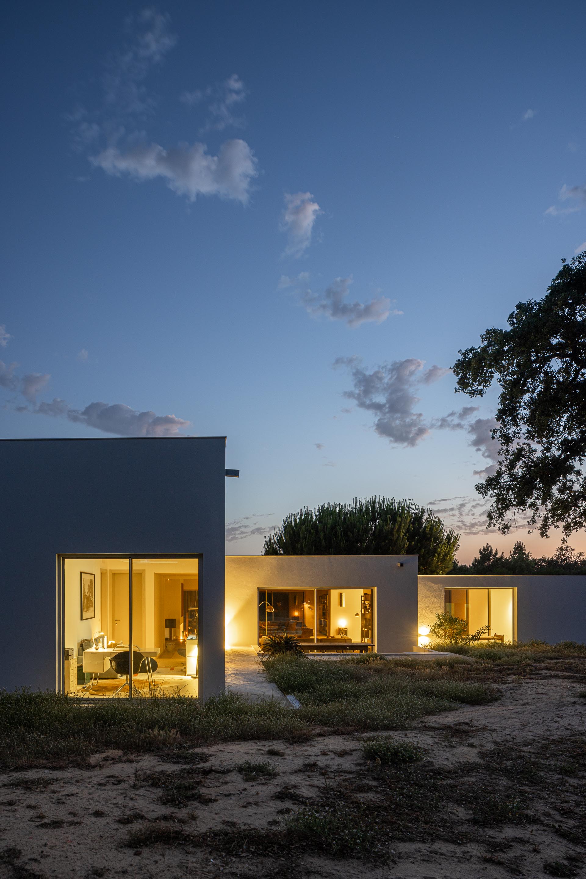 Fachada branca da moradia com vãos envidraçados iluminados, revelando os interiores ao anoitecer — Casa M Ariane fotografia Ivo Tavares