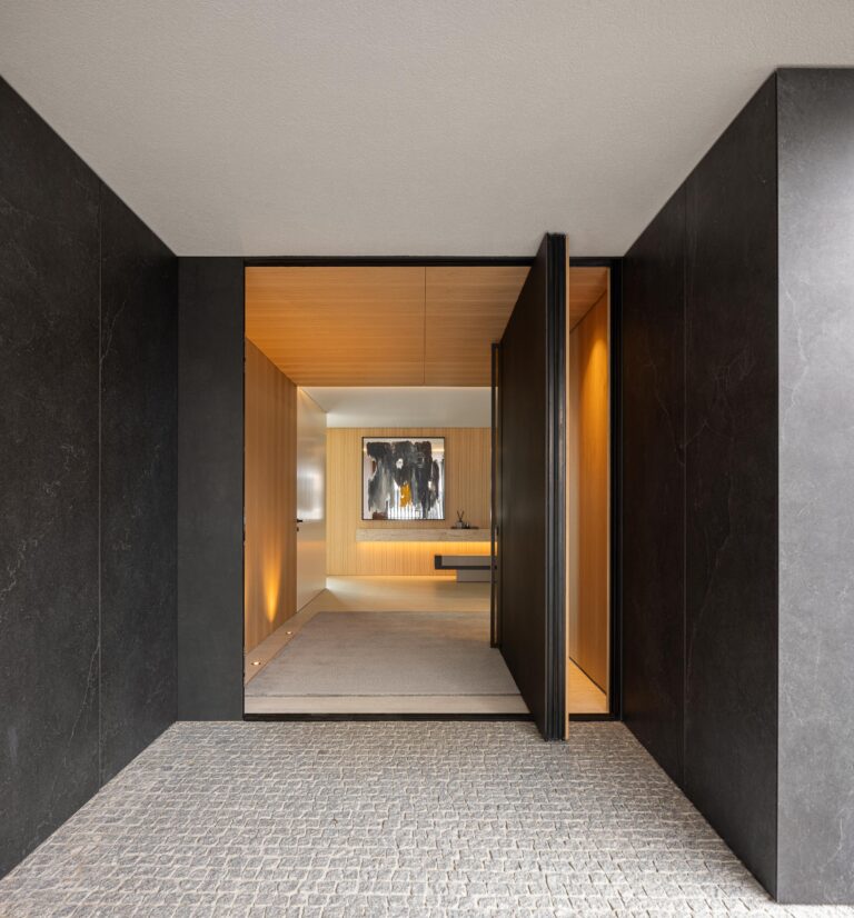 Hall de entrada com pavimento em calçada, paredes em pedra negra e porta pivotante em madeira mel — Casa FJ arquitectura MIL870 + GRAUON fotografia Ivo Tavares