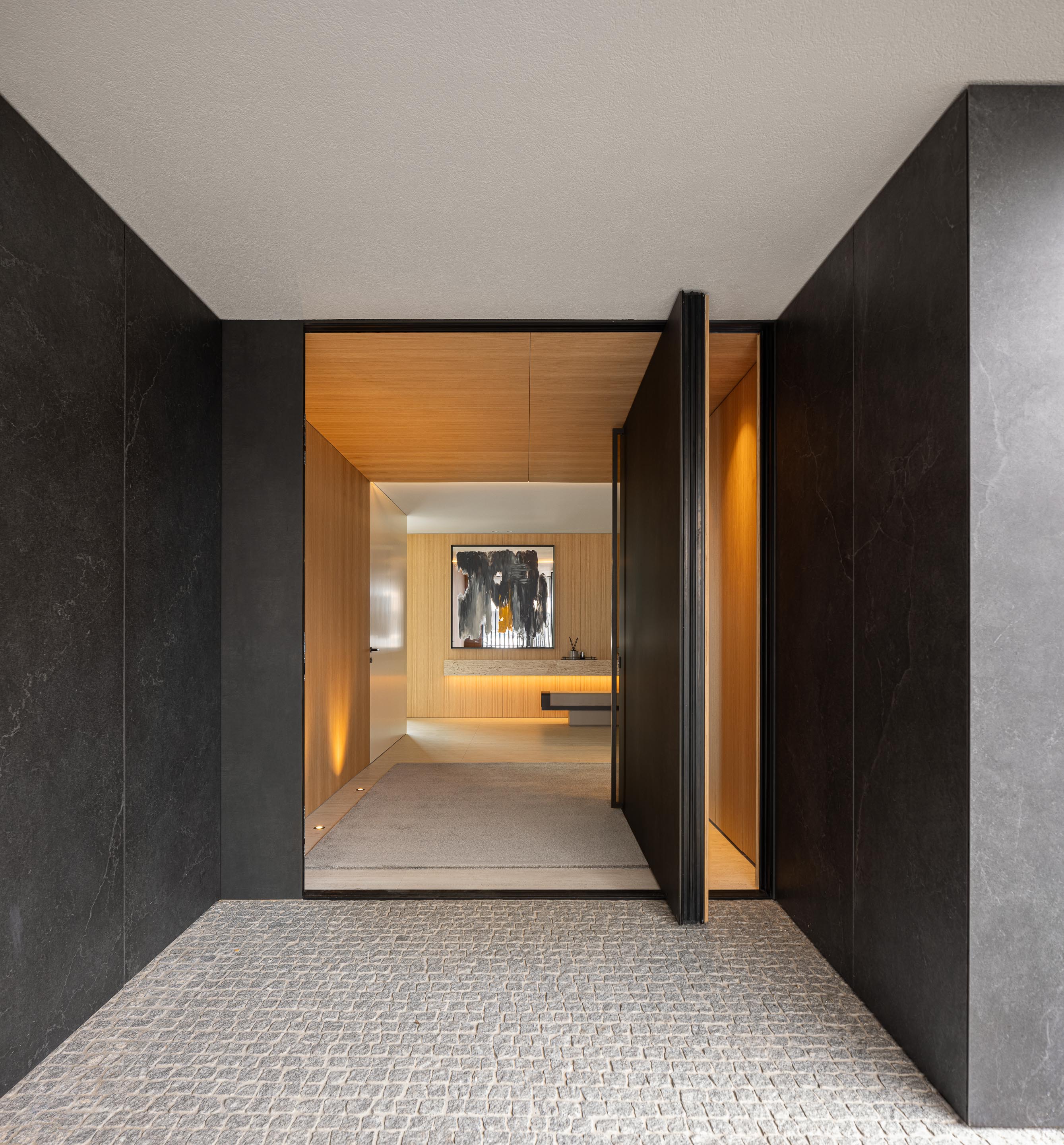 Hall de entrada com pavimento em calçada, paredes em pedra negra e porta pivotante em madeira mel — Casa FJ arquitectura MIL870 + GRAUON fotografia Ivo Tavares