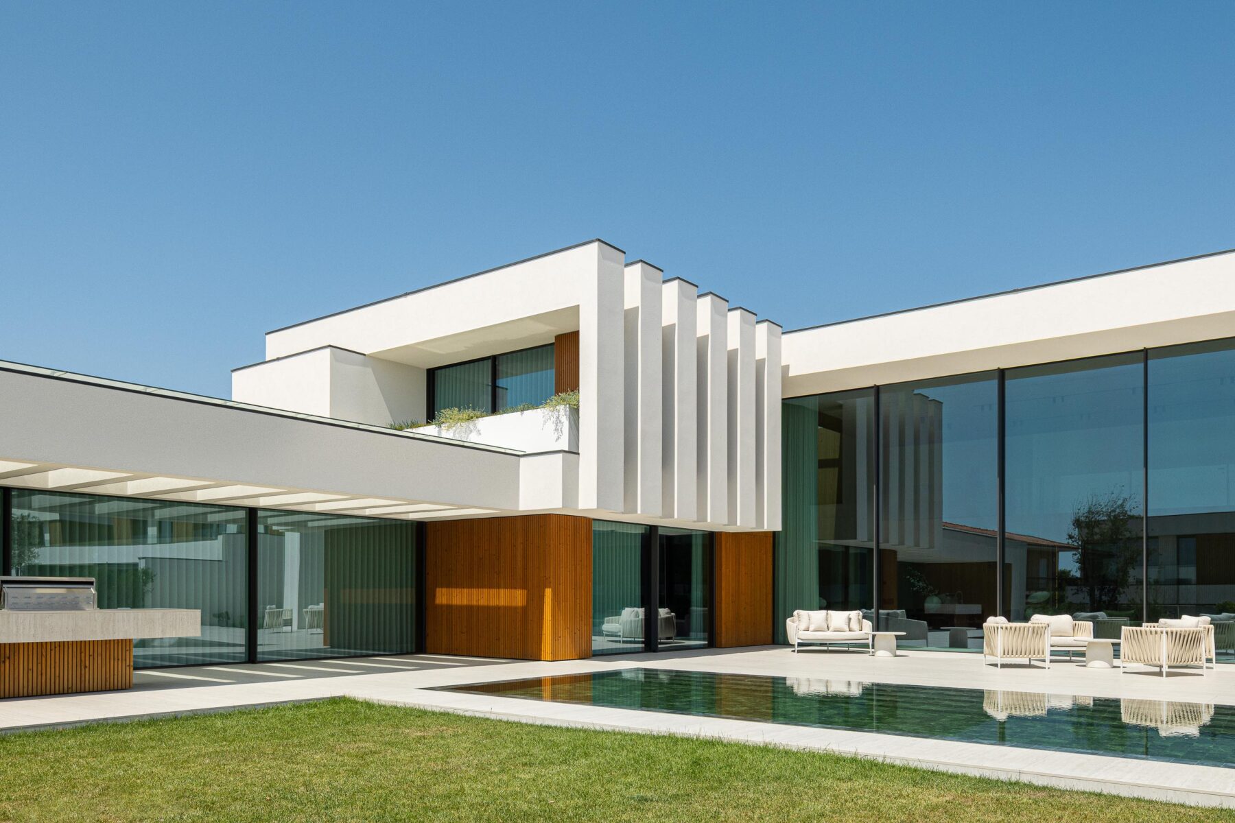 Fachada posterior com volumes brancos, madeira mel e grandes vãos envidraçados sobre a piscina — Casa FJ arquitectura MIL870 + GRAUON fotografia Ivo Tavares