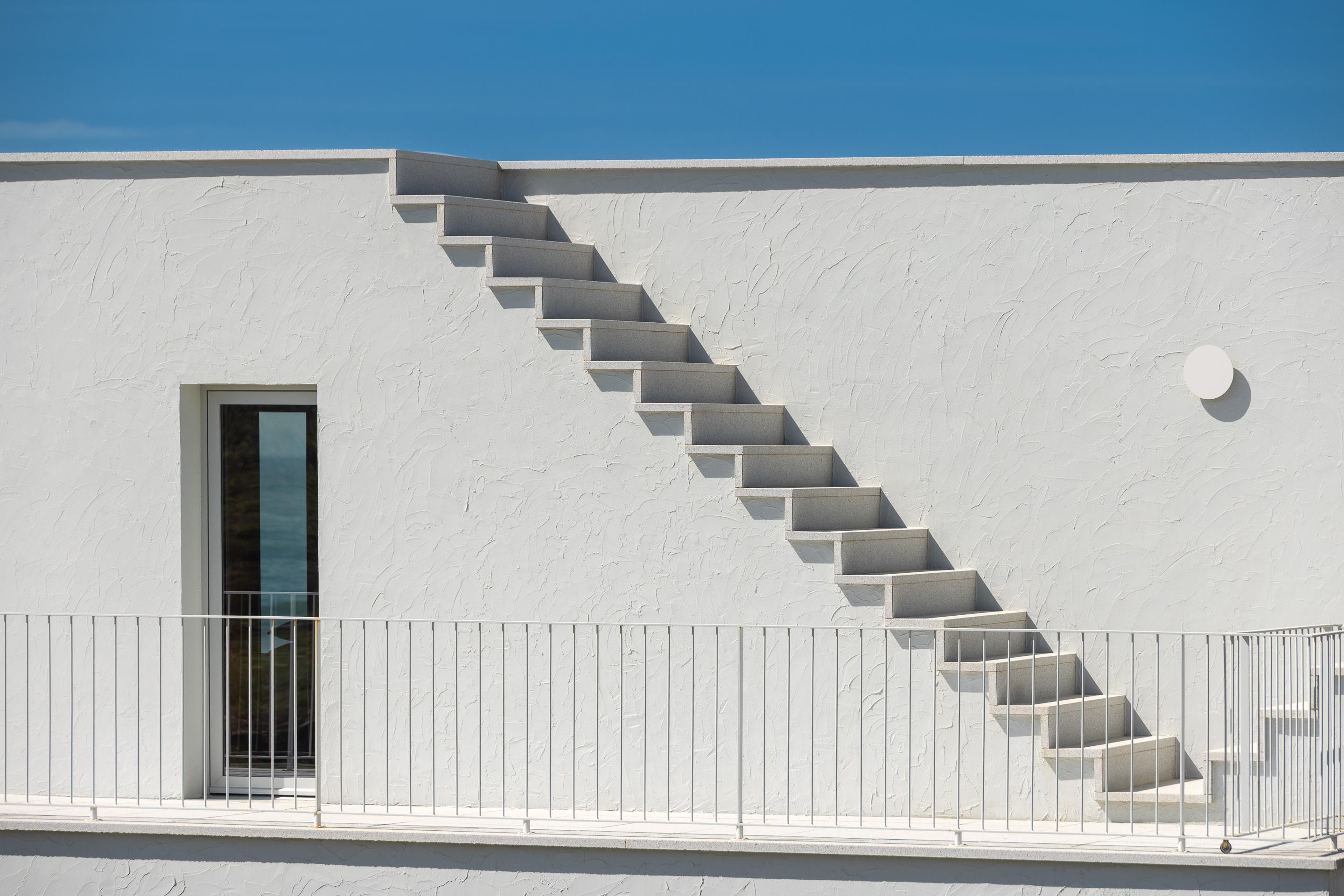 Escadaria exterior em pedra clara sobre fachada branca texturada, com varanda e guarda-corpos — CASA ÂNCORA arquitectura Tiago Sousa Arquitecto fotografia Ivo Tavares