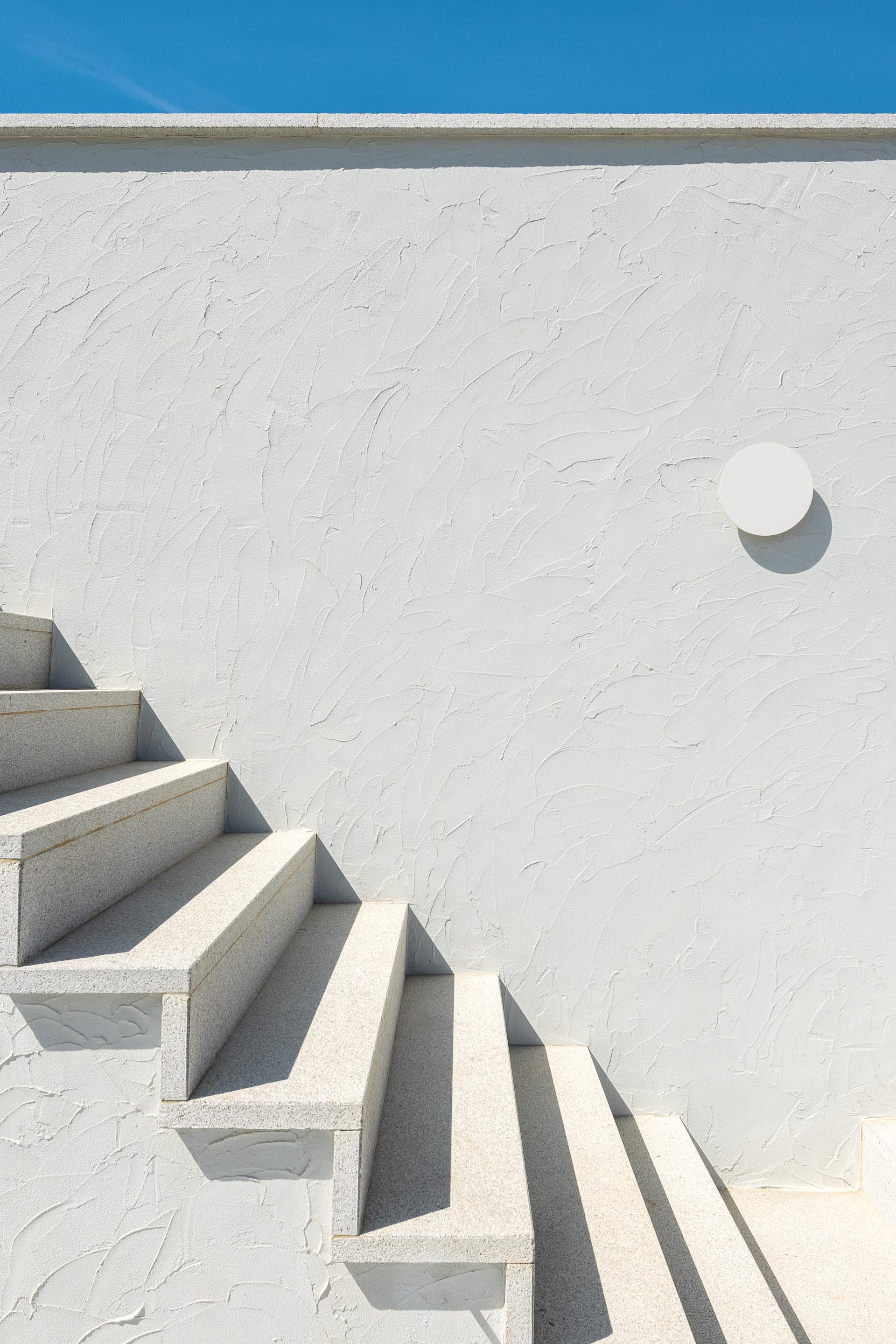 Detalhe da escadaria exterior em pedra clara e fachada branca texturada, com candeeiro circular — CASA ÂNCORA arquitectura Tiago Sousa Arquitecto fotografia Ivo Tavares