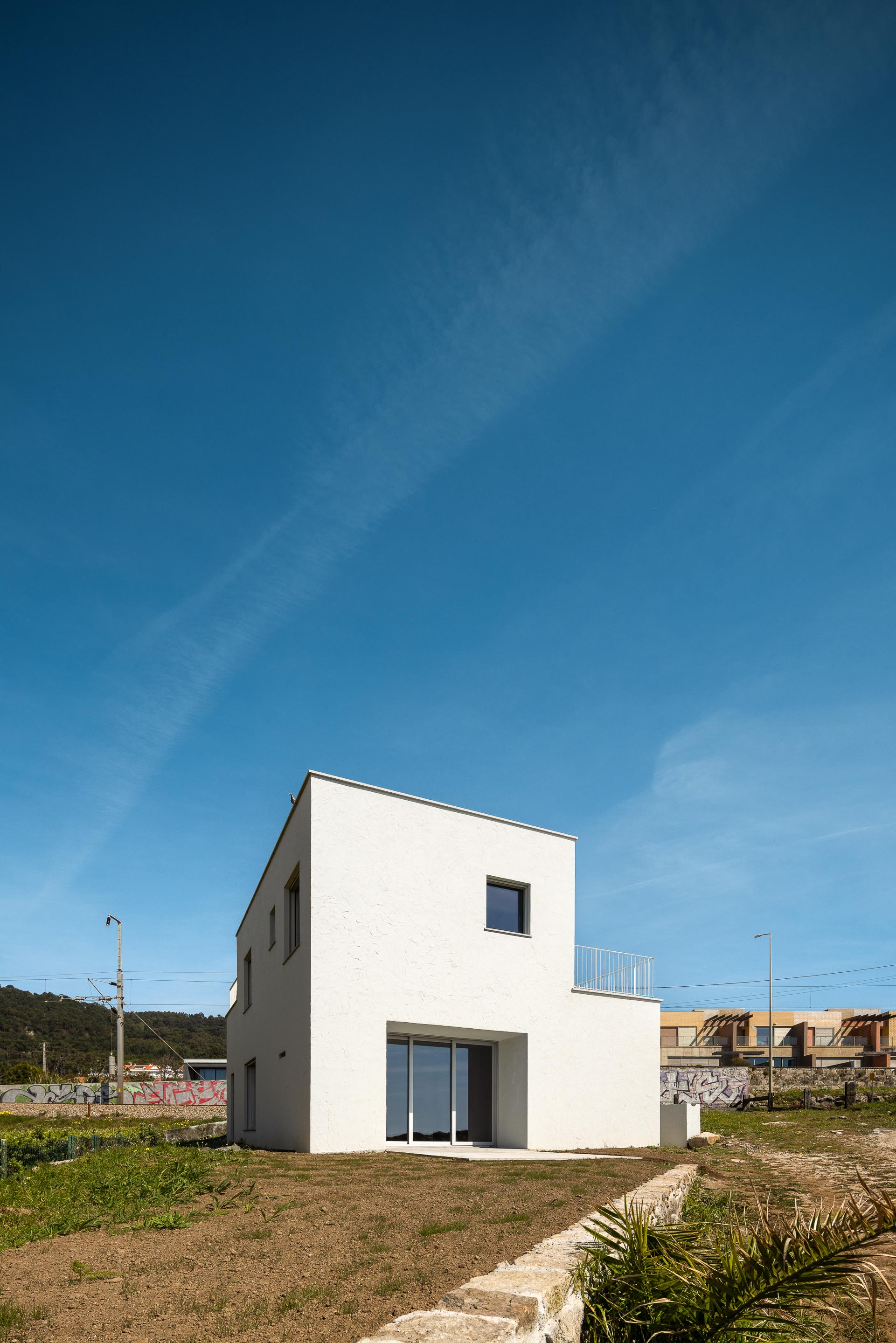 Fachada lateral da moradia branca com cobertura inclinada, vãos e porta de correr, junto à vegetação — CASA ÂNCORA arquitectura Tiago Sousa Arquitecto fotografia Ivo Tavares