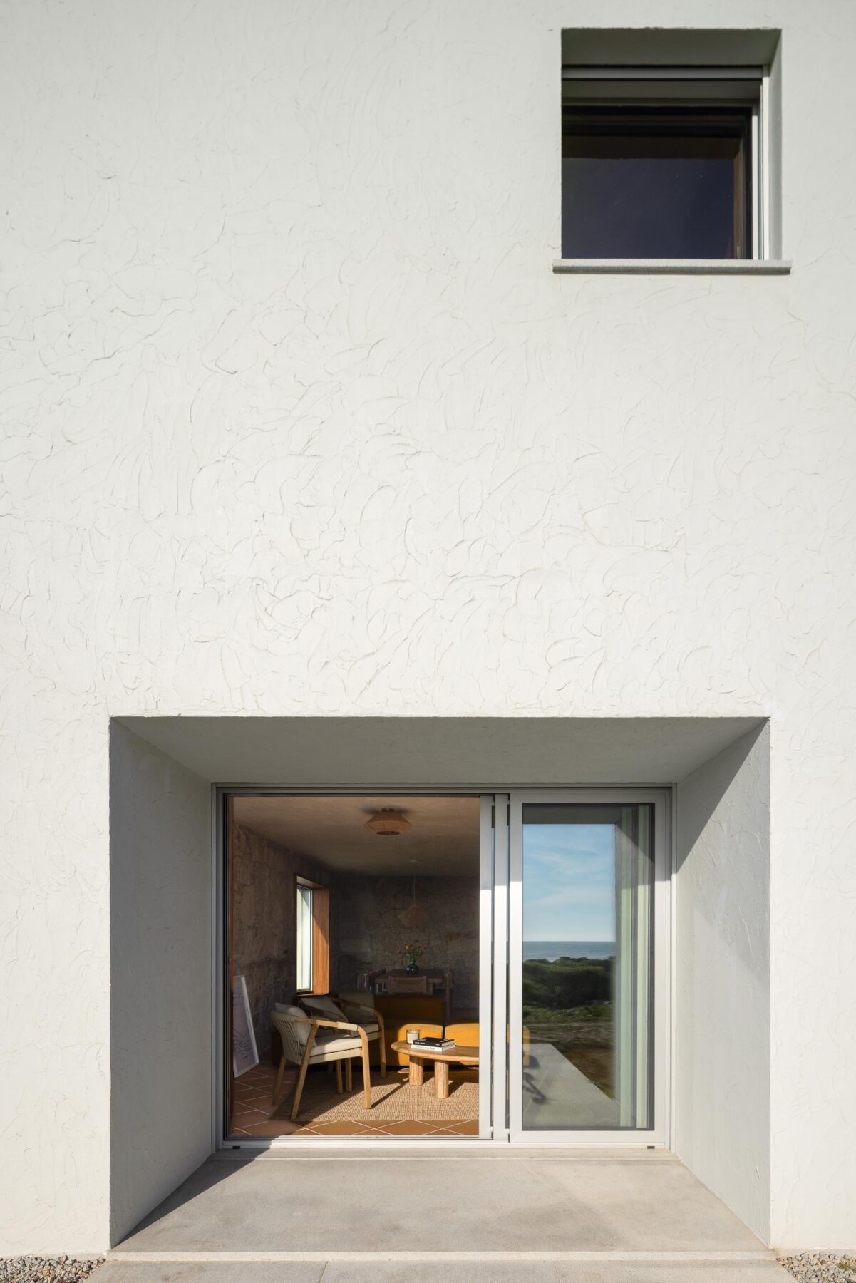 Fachada branca com vão de correr envidraçado, revelando interior com paredes de pedra e pavimento cerâmico — CASA ÂNCORA arquitectura Tiago Sousa Arquitecto fotografia Ivo Tavares