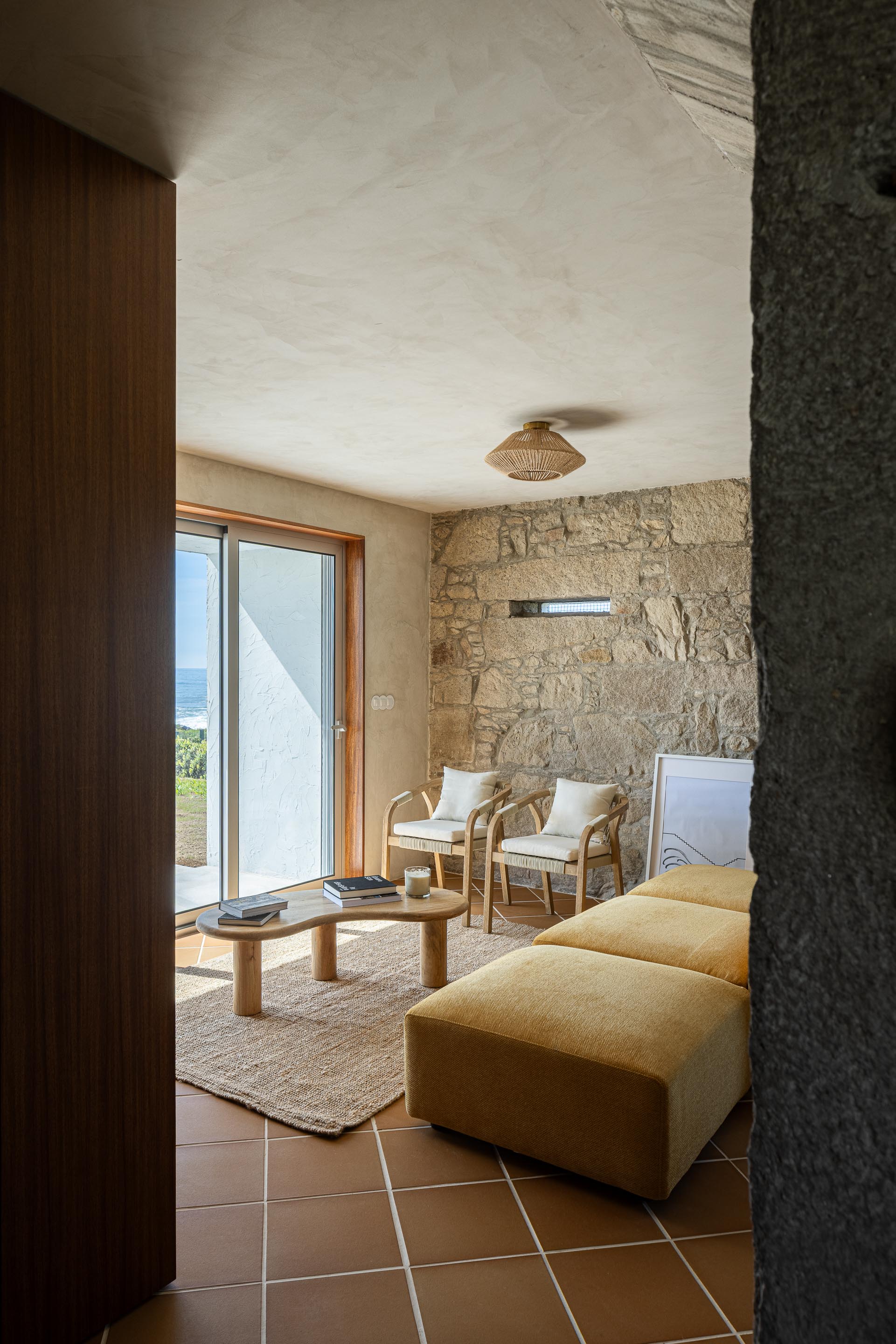 Sala de estar com pavimento cerâmico de barro e parede de pedra, mobiliário em madeira e estofado amarelo — CASA ÂNCORA arquitectura Tiago Sousa Arquitecto fotografia Ivo Tavares
