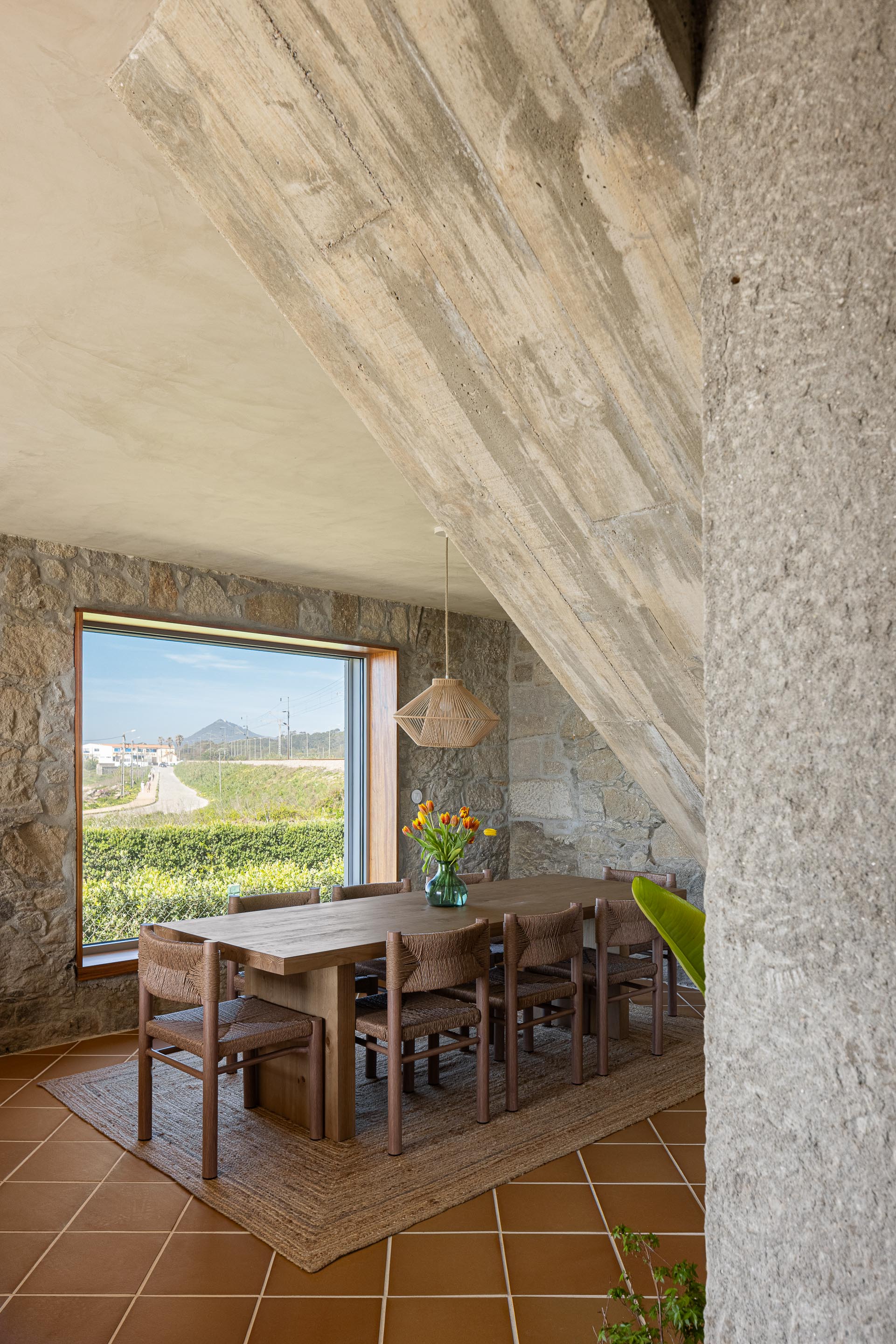 Sala de jantar com parede de pedra, pavimento cerâmico de barro e mesa de madeira, com vista para a paisagem — CASA ÂNCORA arquitectura Tiago Sousa Arquitecto fotografia Ivo Tavares