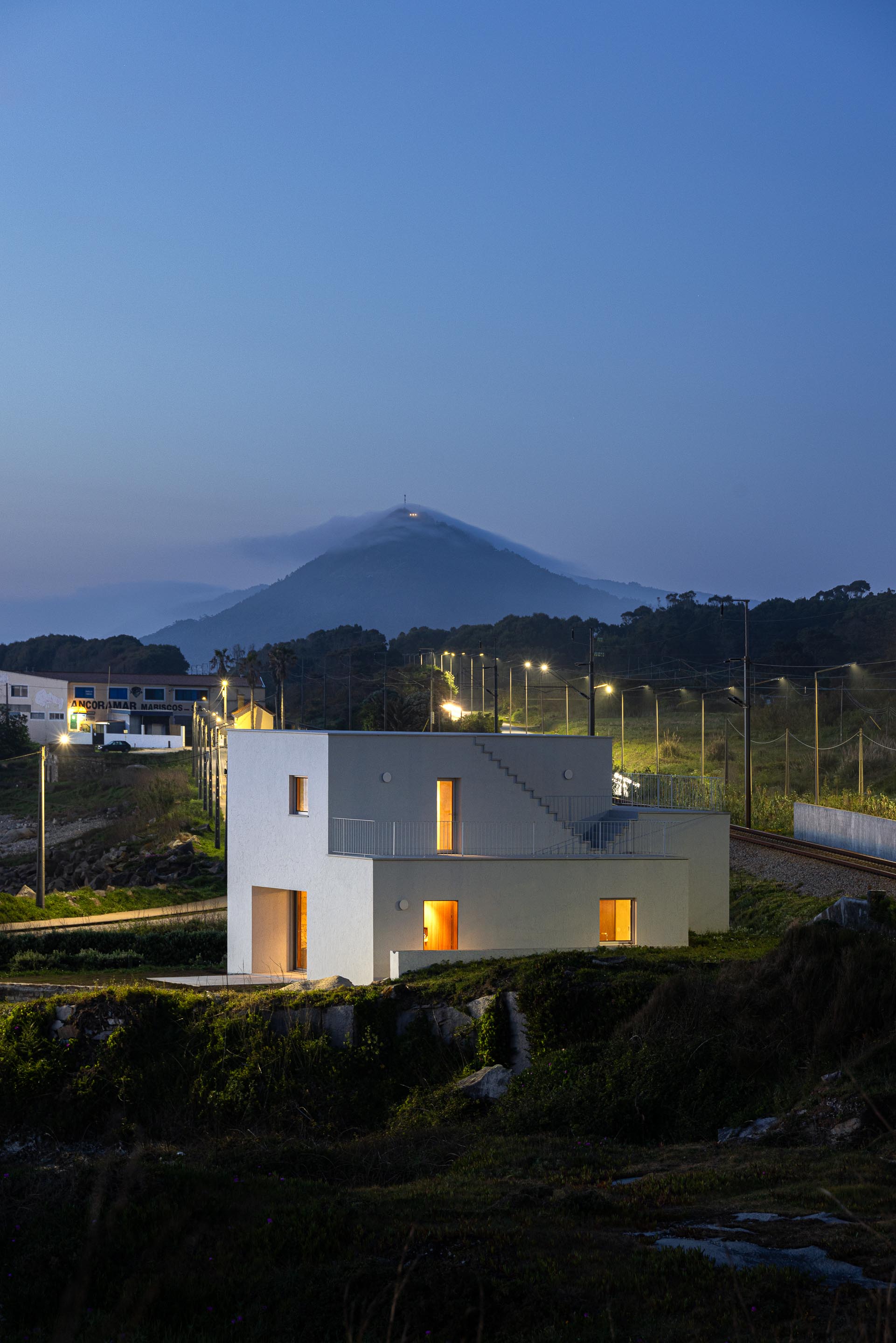 Moradia branca iluminada à noite, com volumes escalonados, inserida na paisagem costeira — CASA ÂNCORA arquitectura Tiago Sousa Arquitecto fotografia Ivo Tavares