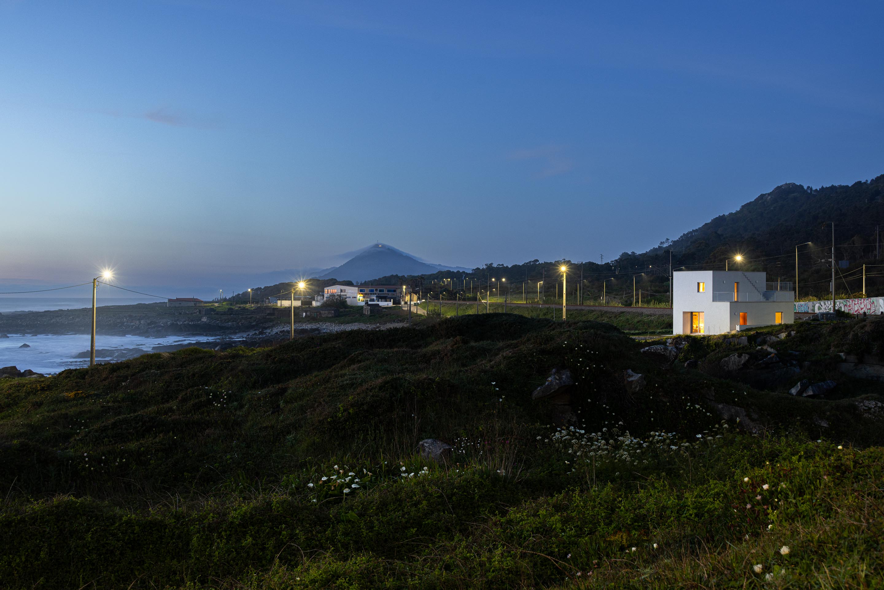 Moradia branca iluminada à noite, integrada na paisagem costeira e montanhosa, sob céu azul escuro — CASA ÂNCORA arquitectura Tiago Sousa Arquitecto fotografia Ivo Tavares