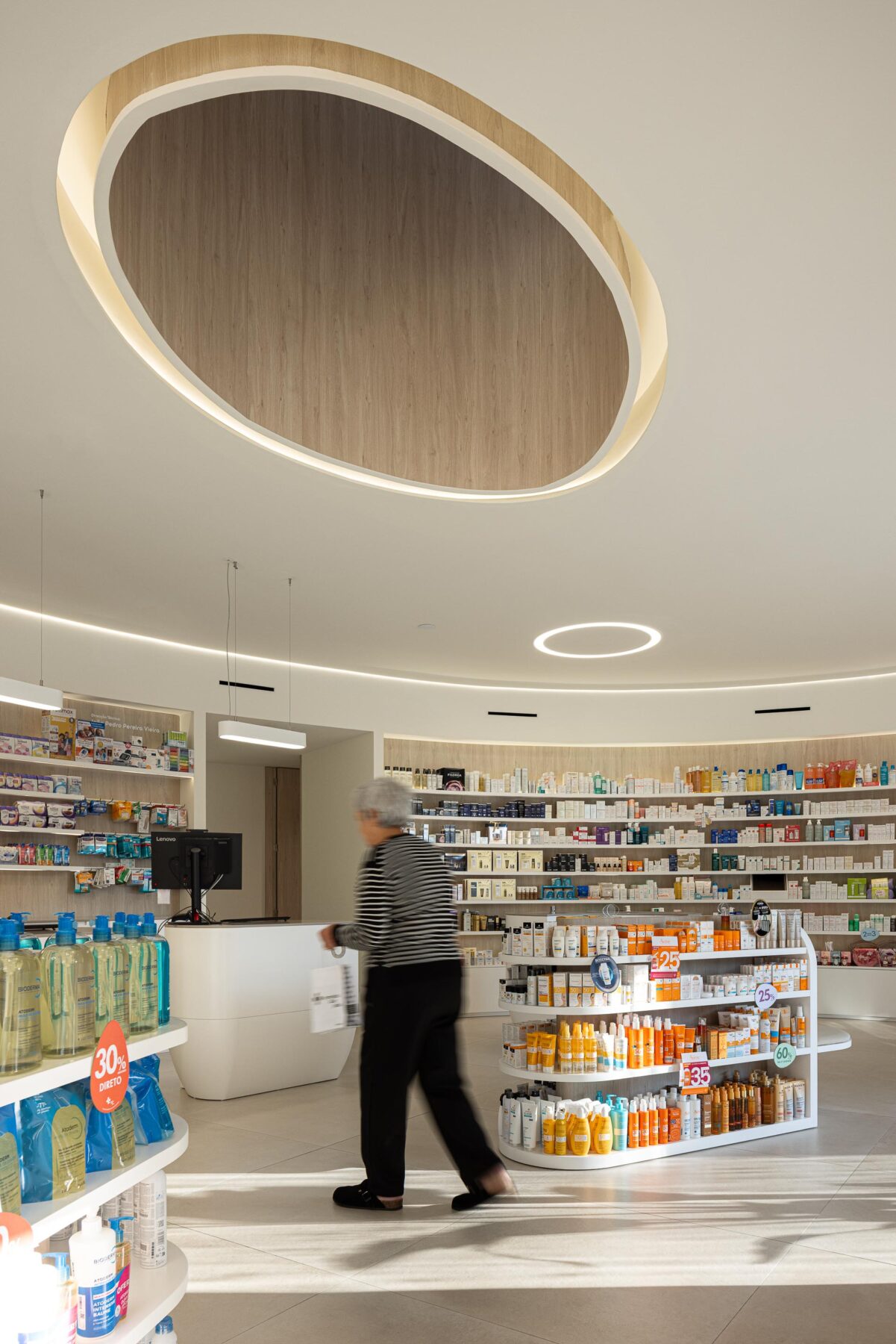 Interior da área de atendimento com clarabóia oval e iluminação zenital, pavimento cerâmico e mobiliário curvo — Farmácia São João arquitectura Tsou Arquitectos fotografia Ivo Tavares