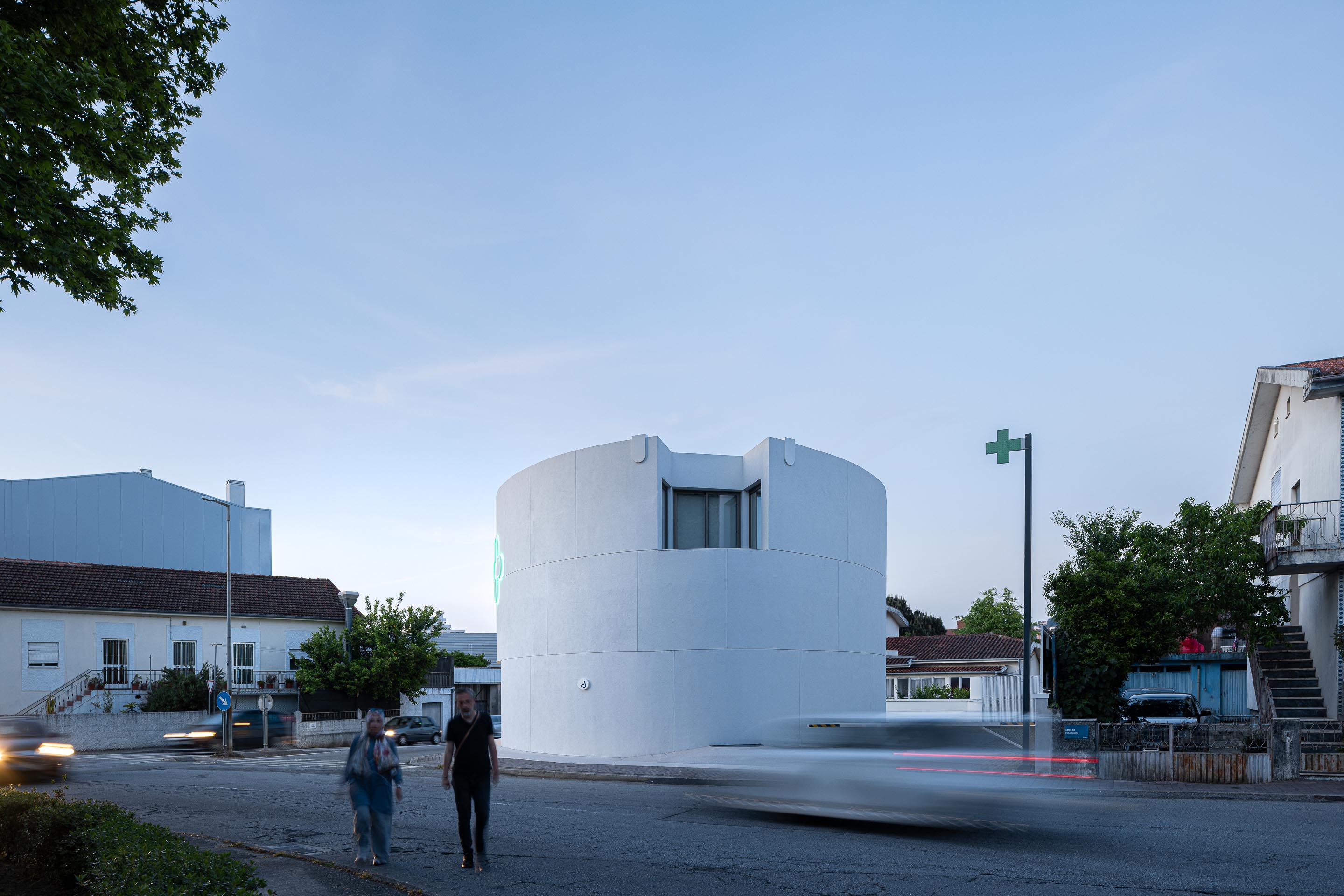 Fachada curva em EPS projetado branco com reclamos luminosos verdes e montras iluminadas ao crepúsculo — Farmácia São João arquitectura Tsou Arquitectos fotografia Ivo Tavares
