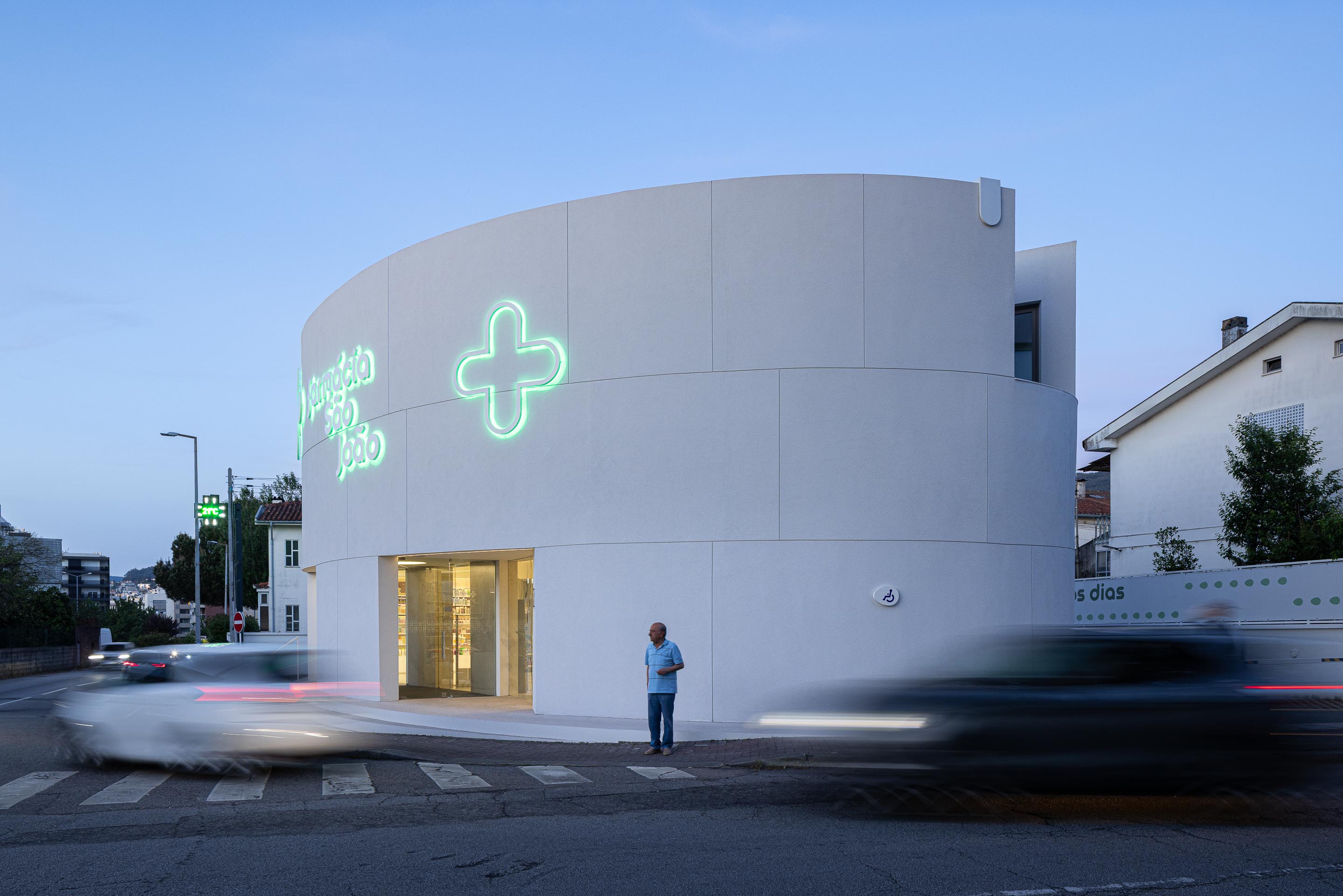 Fachada curva em EPS projetado branco com vão de janela recuado e reclamo luminoso verde, ao crepúsculo — Farmácia São João arquitectura Tsou Arquitectos fotografia Ivo Tavares