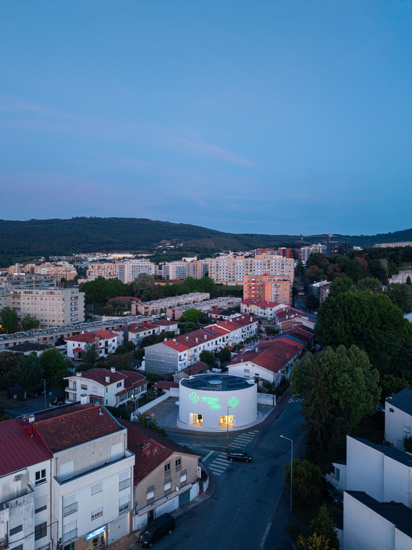 Fachada curva em EPS projetado branco com reclamos luminosos verdes e montras iluminadas, sob luz crepuscular — Farmácia São João arquitectura Tsou Arquitectos fotografia Ivo Tavares