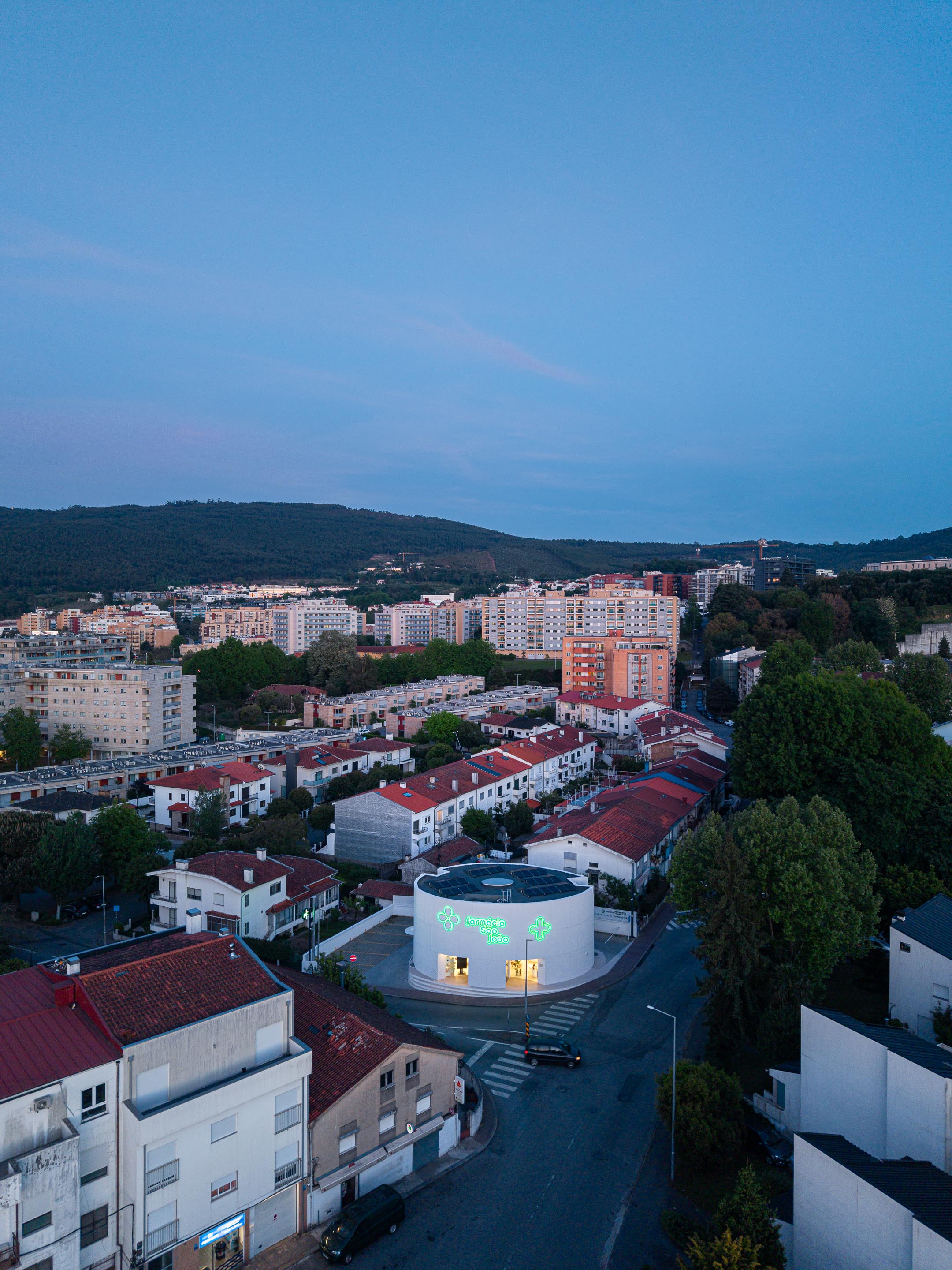 Fachada curva em EPS projetado branco com reclamos luminosos verdes e montras iluminadas, sob luz crepuscular — Farmácia São João arquitectura Tsou Arquitectos fotografia Ivo Tavares