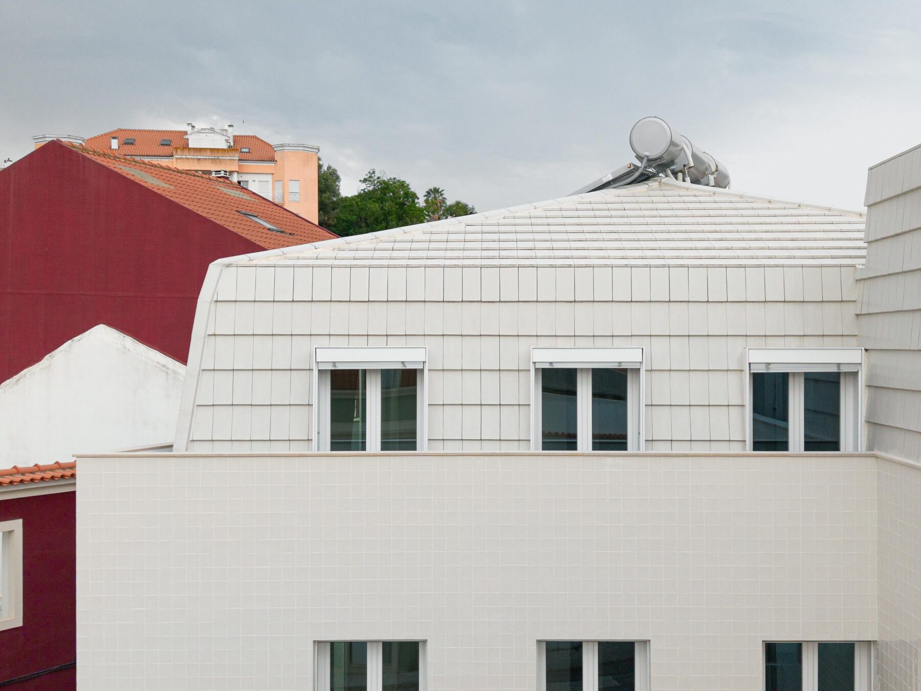 Fachada com revestimento em azulejo branco e cobertura amansardada em telha plana branca. Vãos com caixilharia de PVC branca — Edifício Actriz Virginia arquitectura Adarq – André David Arquitecto fotografia Ivo Tavares