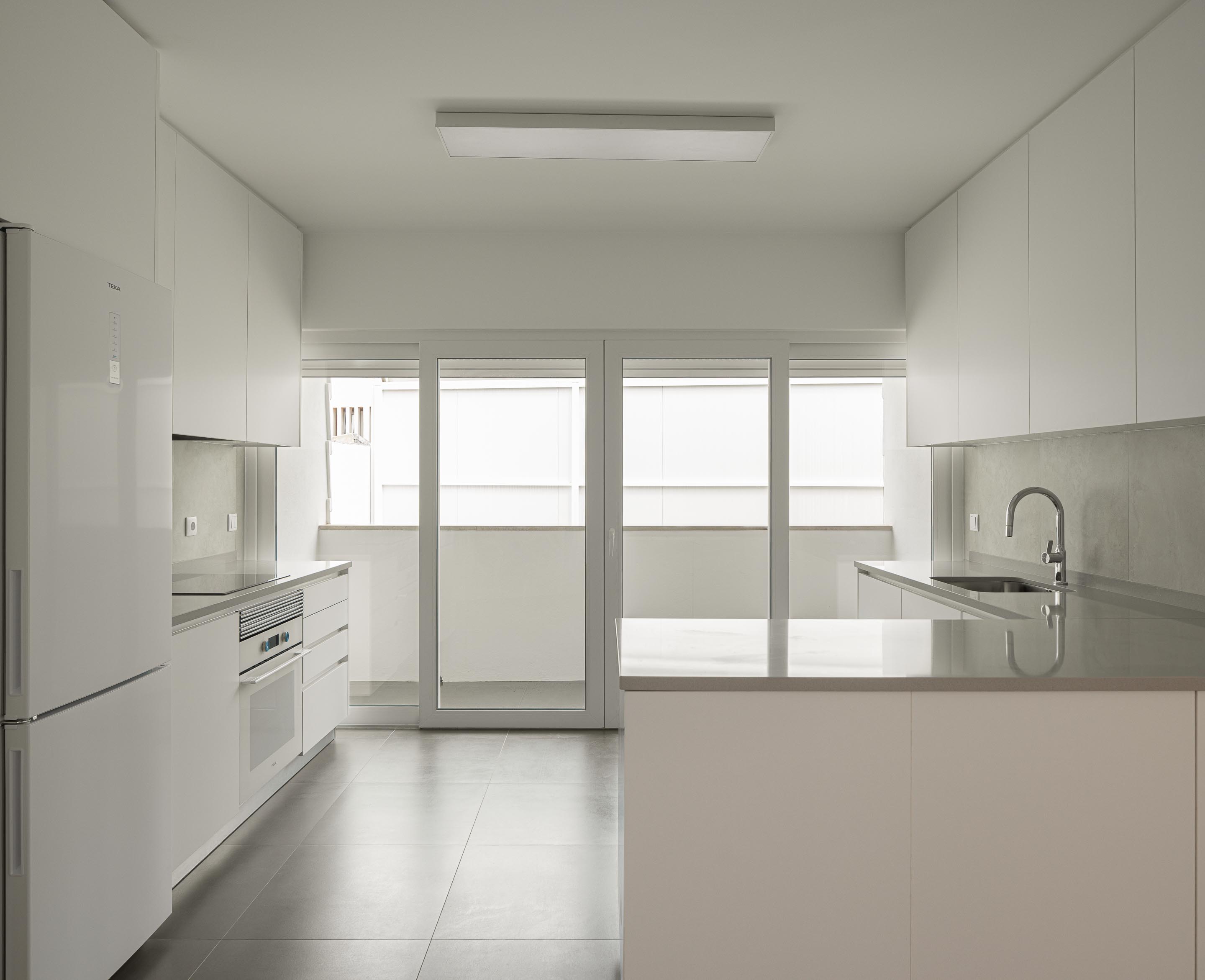 Cozinha com mobiliário branco minimalista, pavimento em cerâmica cinzenta e bancadas claras. Vão envidraçado para o exterior — Edifício Actriz Virginia arquitectura Adarq – André David Arquitecto fotografia Ivo Tavares