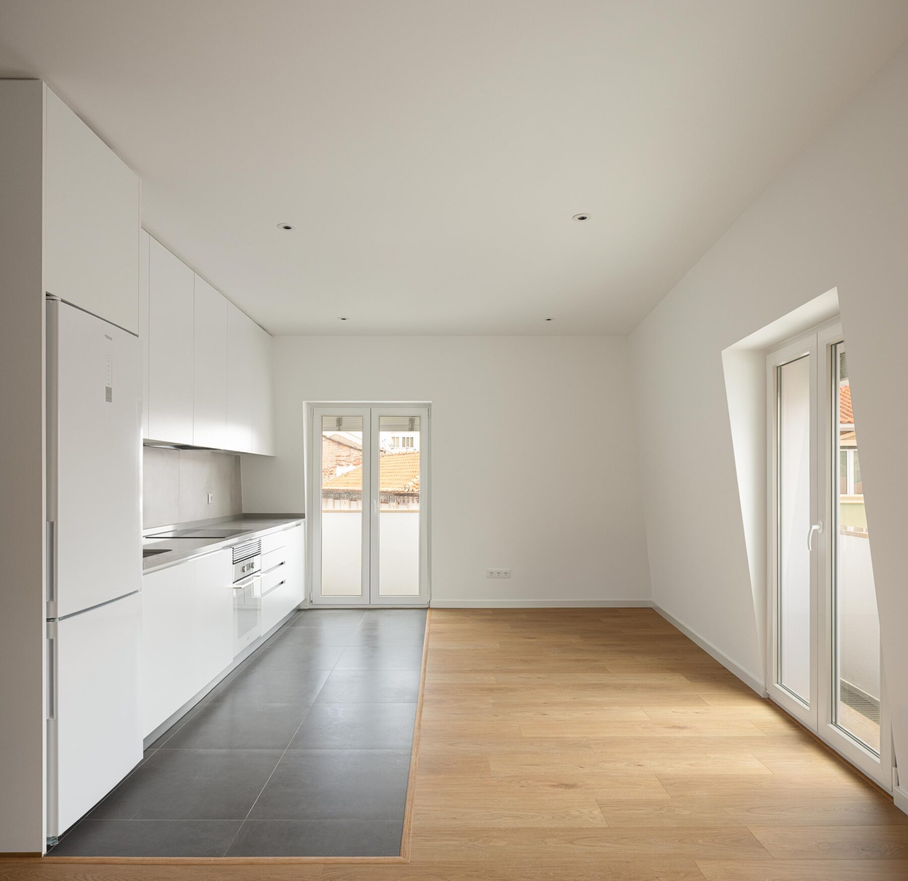 Cozinha e sala em open space com pavimento misto: cerâmica cinzenta e madeira de carvalho clara. Mobiliário de cozinha branco — Edifício Actriz Virginia arquitectura Adarq – André David Arquitecto fotografia Ivo Tavares