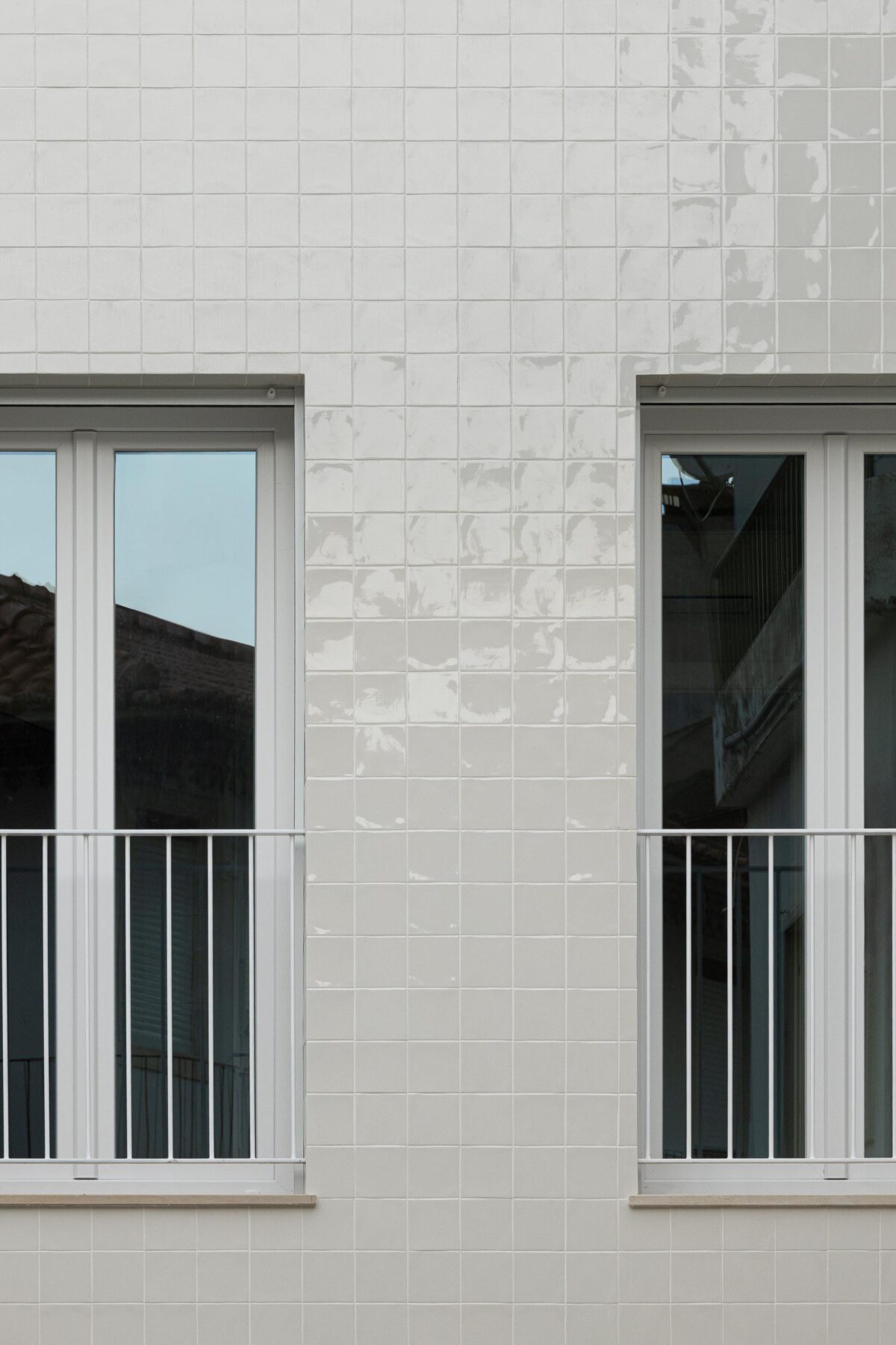 Dois vãos na fachada em azulejo branco, com caixilharia de PVC branca e guarda-corpos metálicos. Reflexos no vidro — Edifício Actriz Virginia arquitectura Adarq – André David Arquitecto fotografia Ivo Tavares