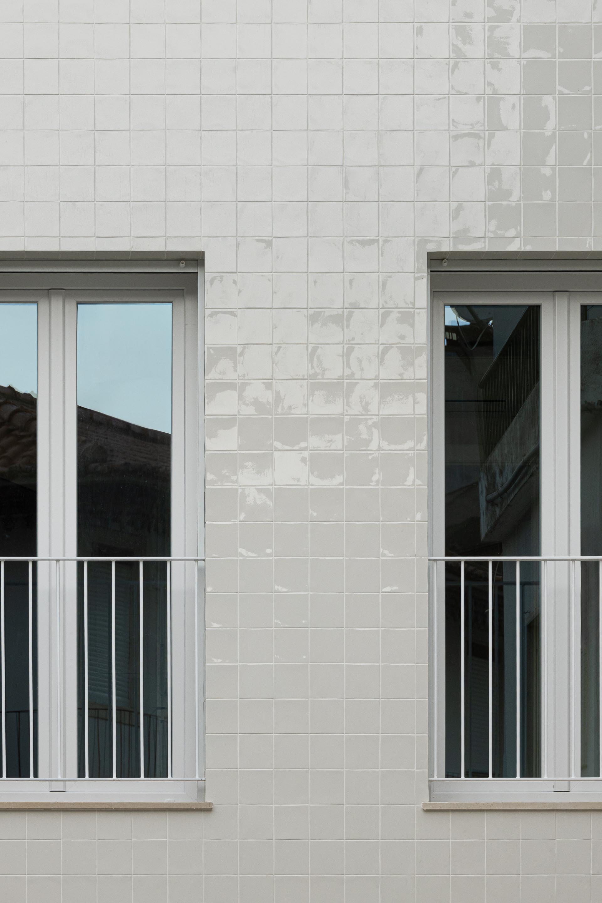Dois vãos na fachada em azulejo branco, com caixilharia de PVC branca e guarda-corpos metálicos. Reflexos no vidro — Edifício Actriz Virginia arquitectura Adarq – André David Arquitecto fotografia Ivo Tavares