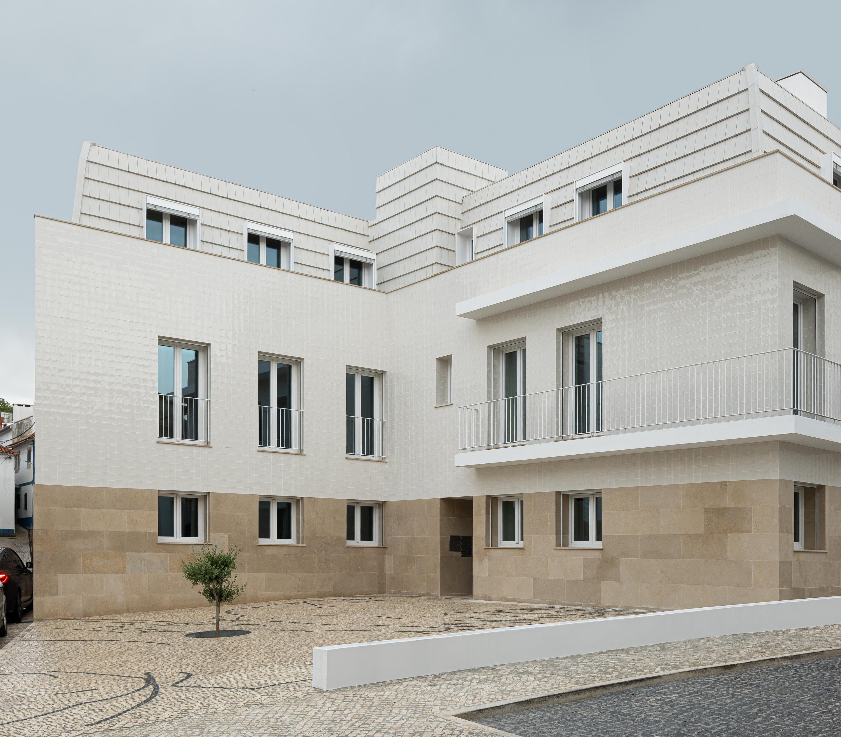 Edifício em L com base em pedra calcária, fachada em azulejo branco e cobertura amansardada. Calçada portuguesa com árvore — Edifício Actriz Virginia arquitectura Adarq – André David Arquitecto fotografia Ivo Tavares