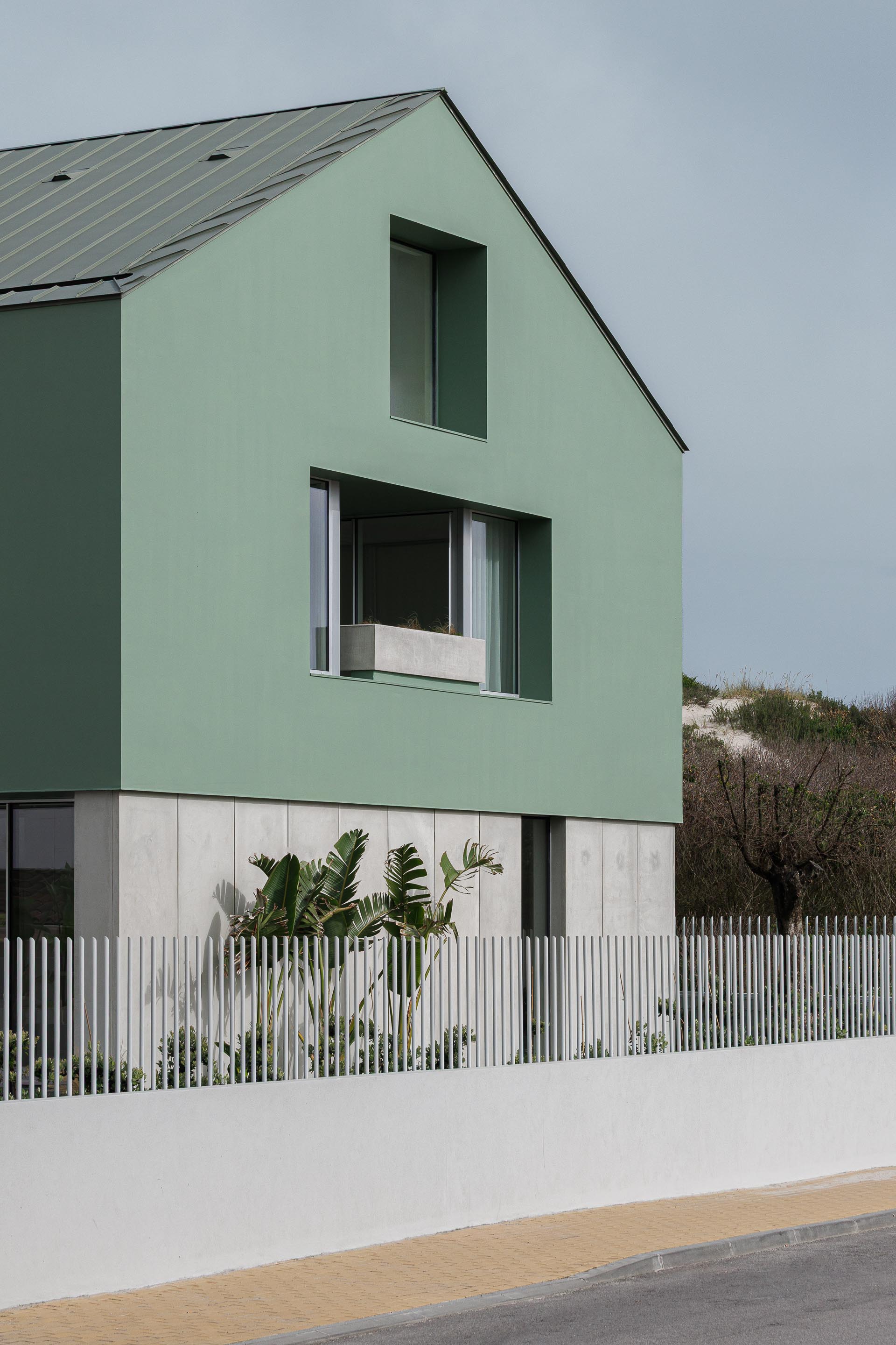 Moradia de volume compacto verde com cobertura inclinada em zinco, embasamento em betão aparente e duna ao fundo — Casa Verde arquitectura M2 Senos Arquitectos fotografia Ivo Tavares