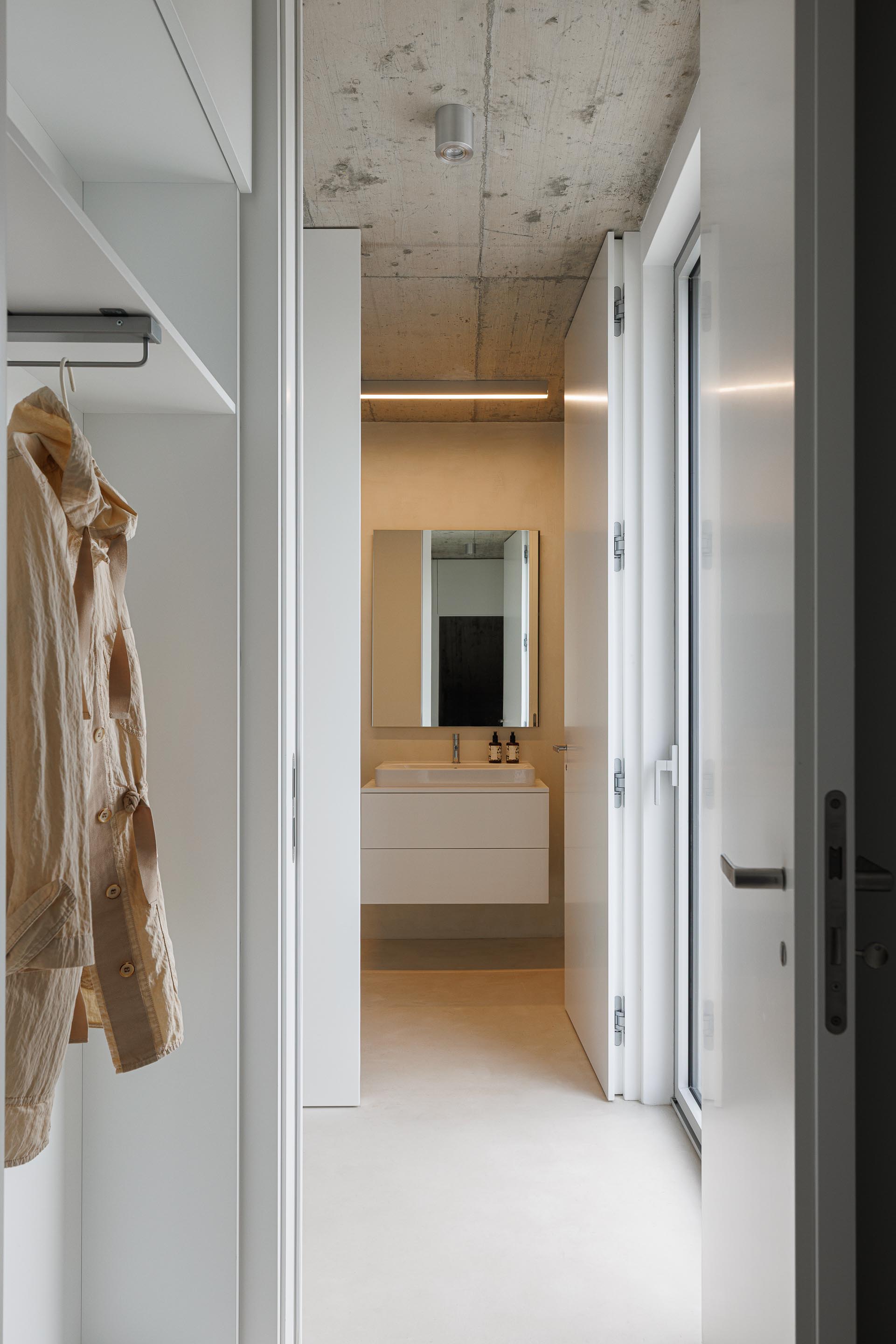Corredor com armário embutido branco e pavimento claro, conduzindo a uma casa de banho com tecto em betão aparente — Casa Verde arquitectura M2 Senos Arquitectos fotografia Ivo Tavares