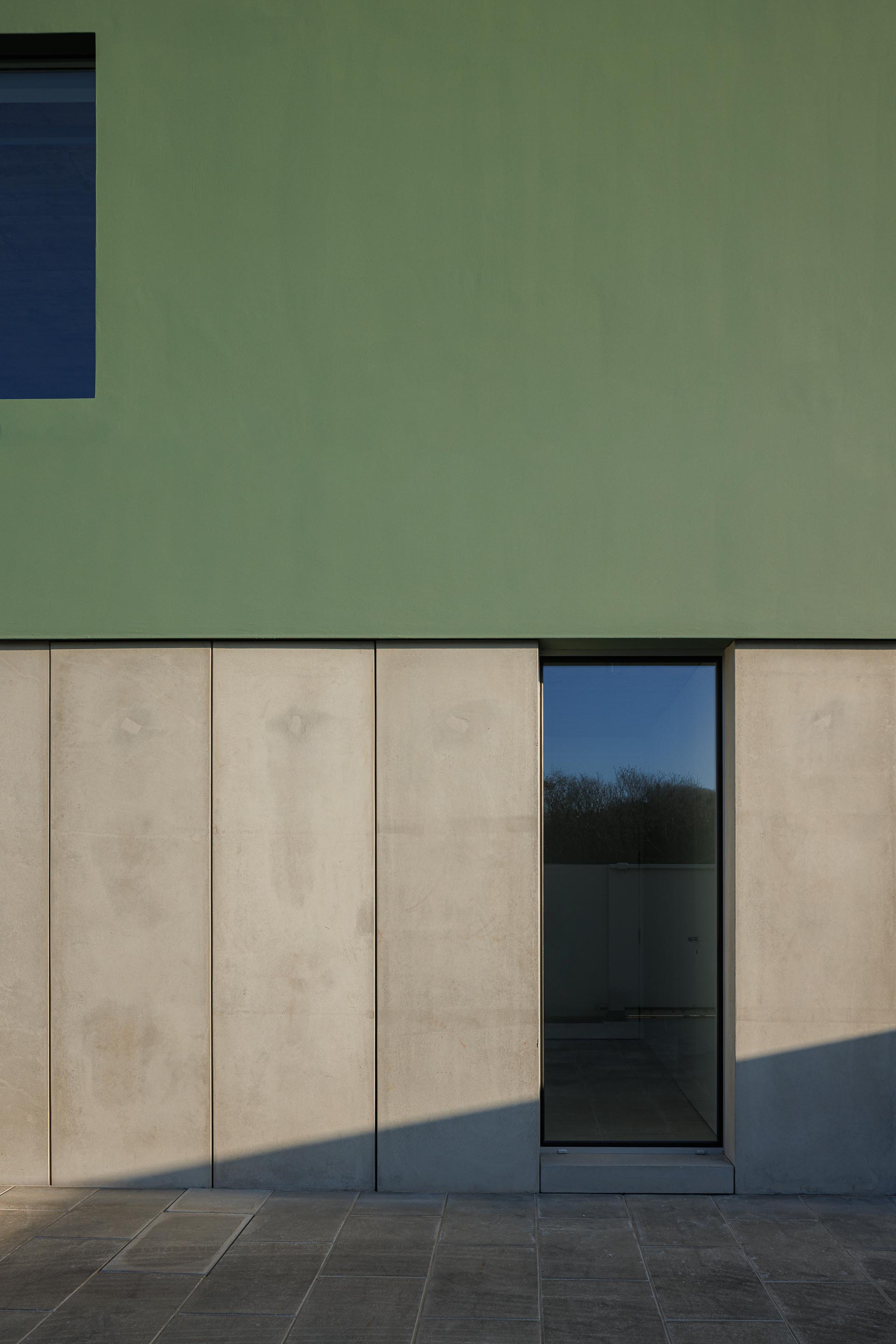 Detalhe da fachada com embasamento em betão aparente e volume superior verde, com porta envidraçada e luz solar — Casa Verde arquitectura M2 Senos Arquitectos fotografia Ivo Tavares