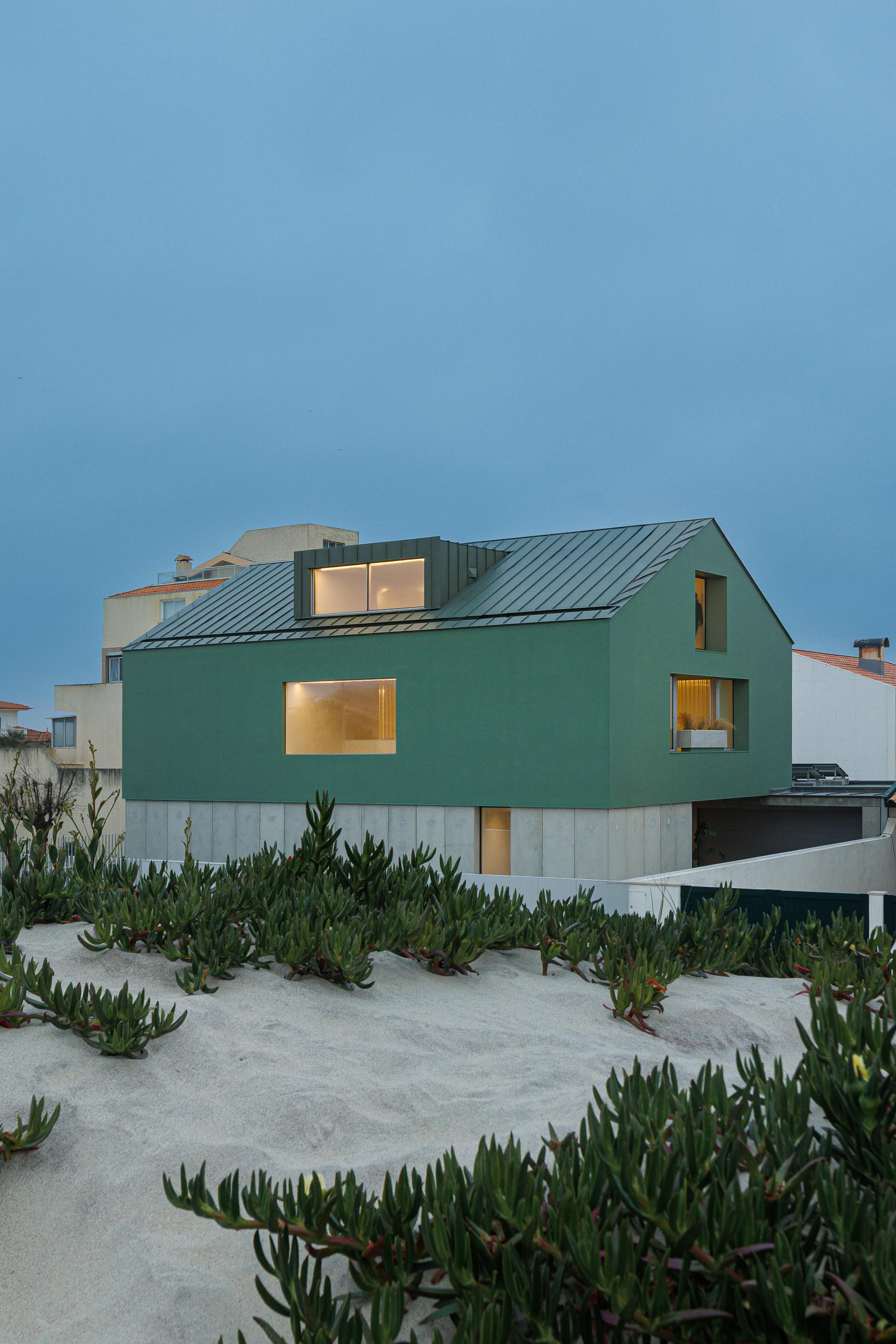 Moradia de volume compacto verde com cobertura inclinada e mansarda, sobre embasamento em betão, integrada na duna ao entardecer — Casa Verde arquitectura M2 Senos Arquitectos fotografia Ivo Tavares