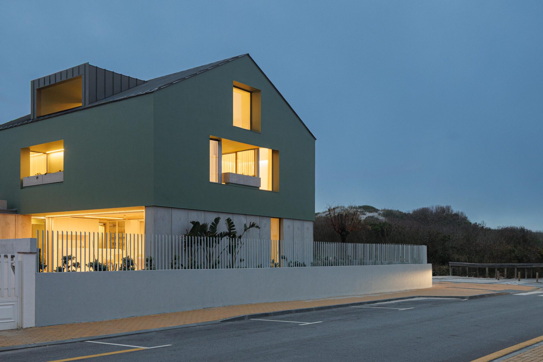Moradia de volume compacto verde com embasamento em betão aparente, vãos iluminados e mansardas, ao entardecer — Casa Verde arquitectura M2 Senos Arquitectos fotografia Ivo Tavares