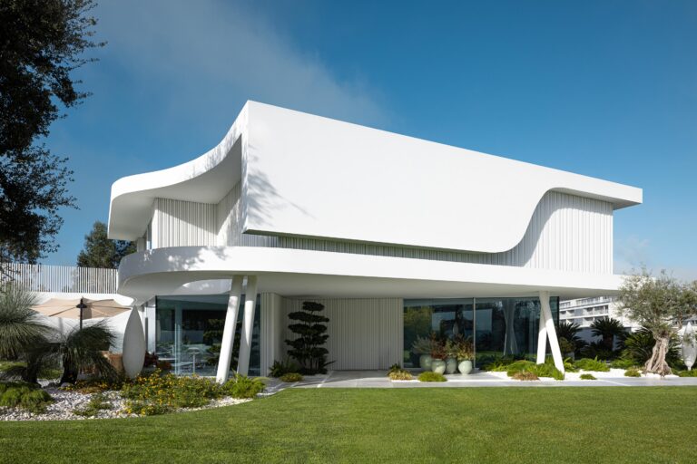 Moradia de volumes brancos curvos, com fachada envidraçada no rés-do-chão e colunas inclinadas, inserida em jardim verde — RiscoWhite arquitectura Risco Singular – Arquitectura, Lda. fotografia Ivo Tavares