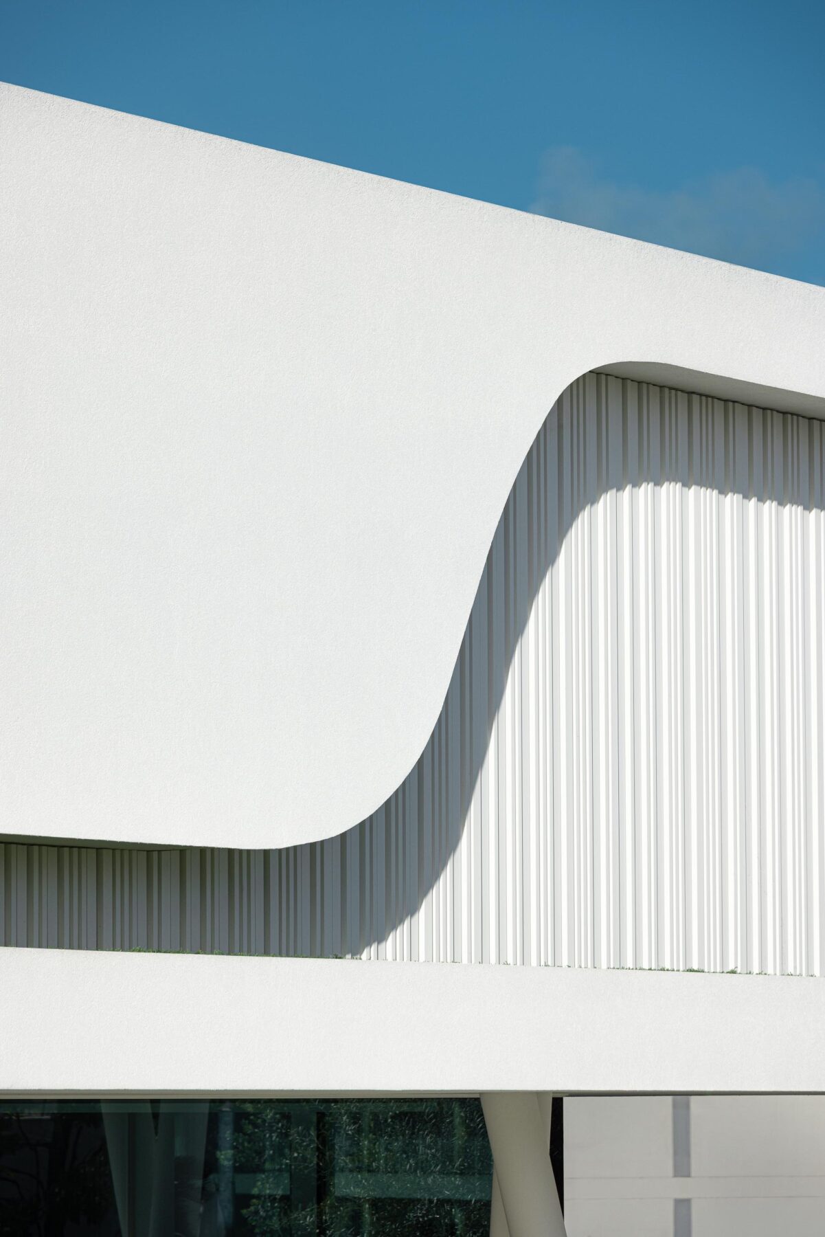 Fachada curva em branco mate com painéis verticais ripados, criando um jogo de luz e sombra dinâmico — RiscoWhite arquitectura Risco Singular – Arquitectura, Lda. fotografia Ivo Tavares