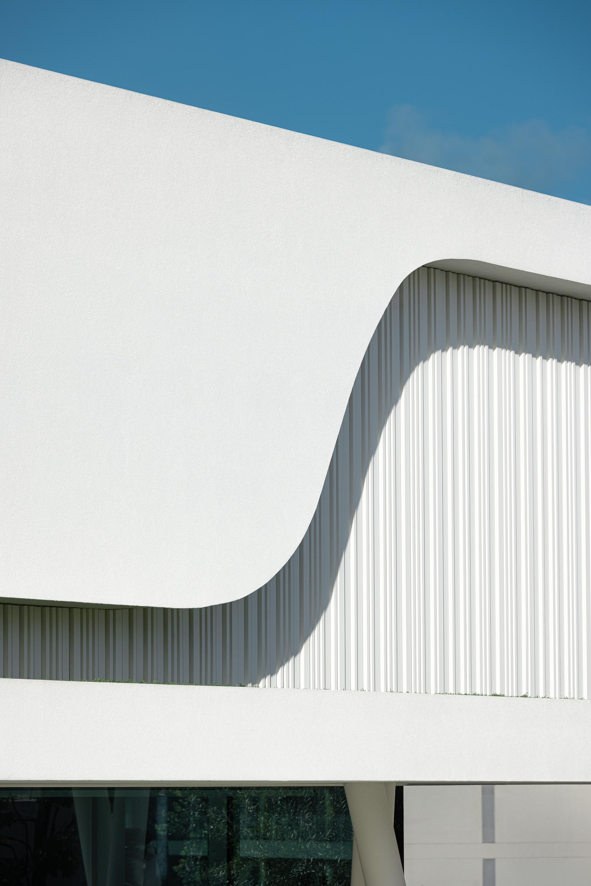 Fachada curva em branco mate com painéis verticais ripados, criando um jogo de luz e sombra dinâmico — RiscoWhite arquitectura Risco Singular – Arquitectura, Lda. fotografia Ivo Tavares