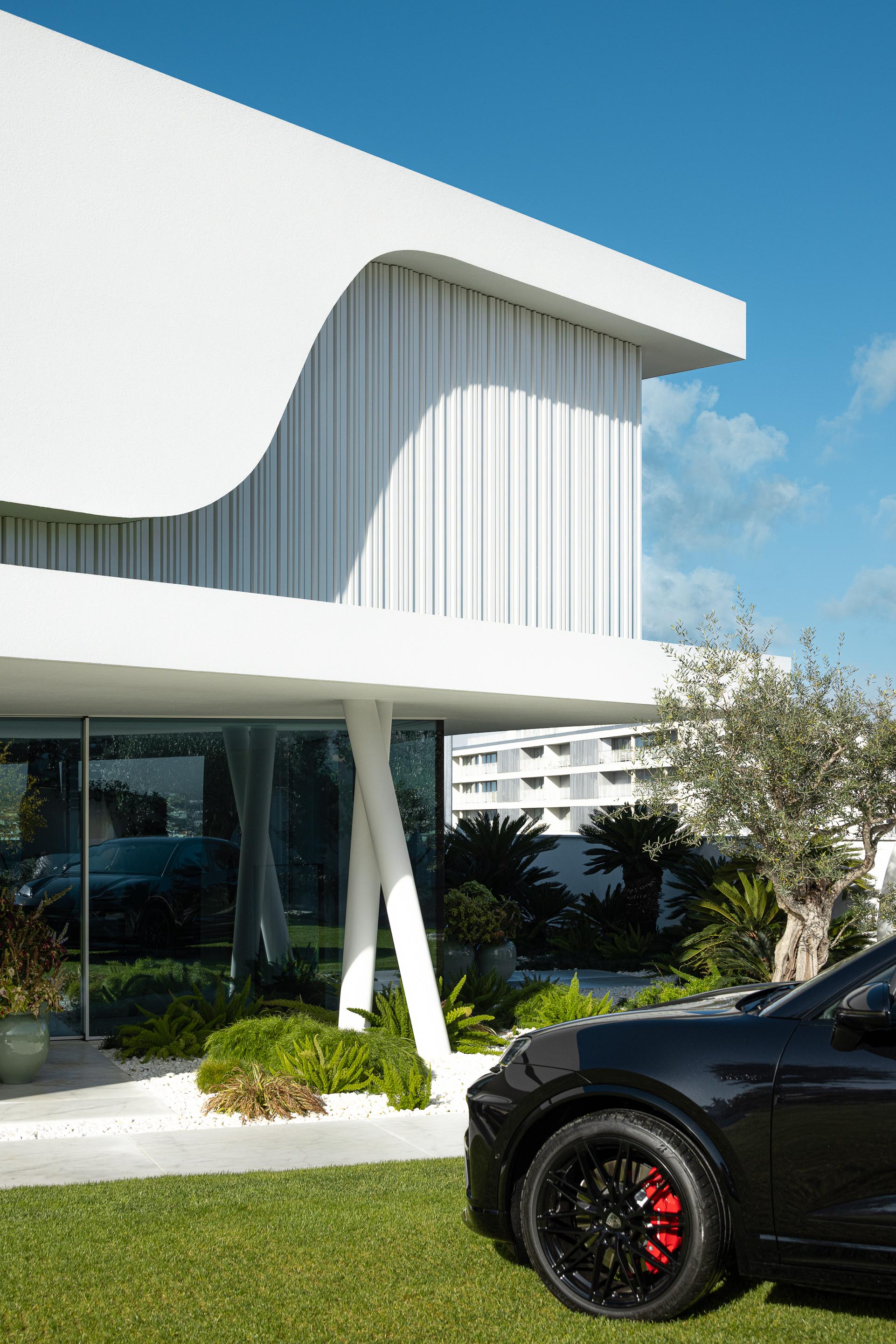Fachada principal da moradia com volumes brancos curvos, vãos envidraçados e colunas inclinadas, com carro estacionado — RiscoWhite arquitectura Risco Singular – Arquitectura, Lda. fotografia Ivo Tavares