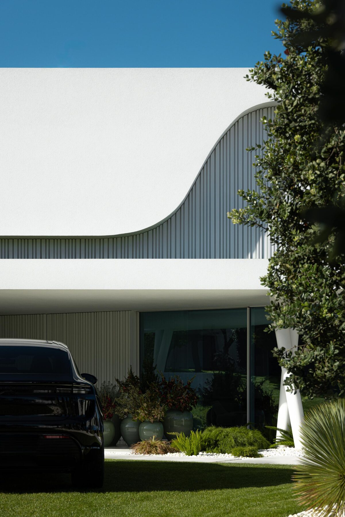Detalhe da fachada branca com elemento curvo e ripado vertical, refletindo a luz e a vegetação circundante — RiscoWhite arquitectura Risco Singular – Arquitectura, Lda. fotografia Ivo Tavares