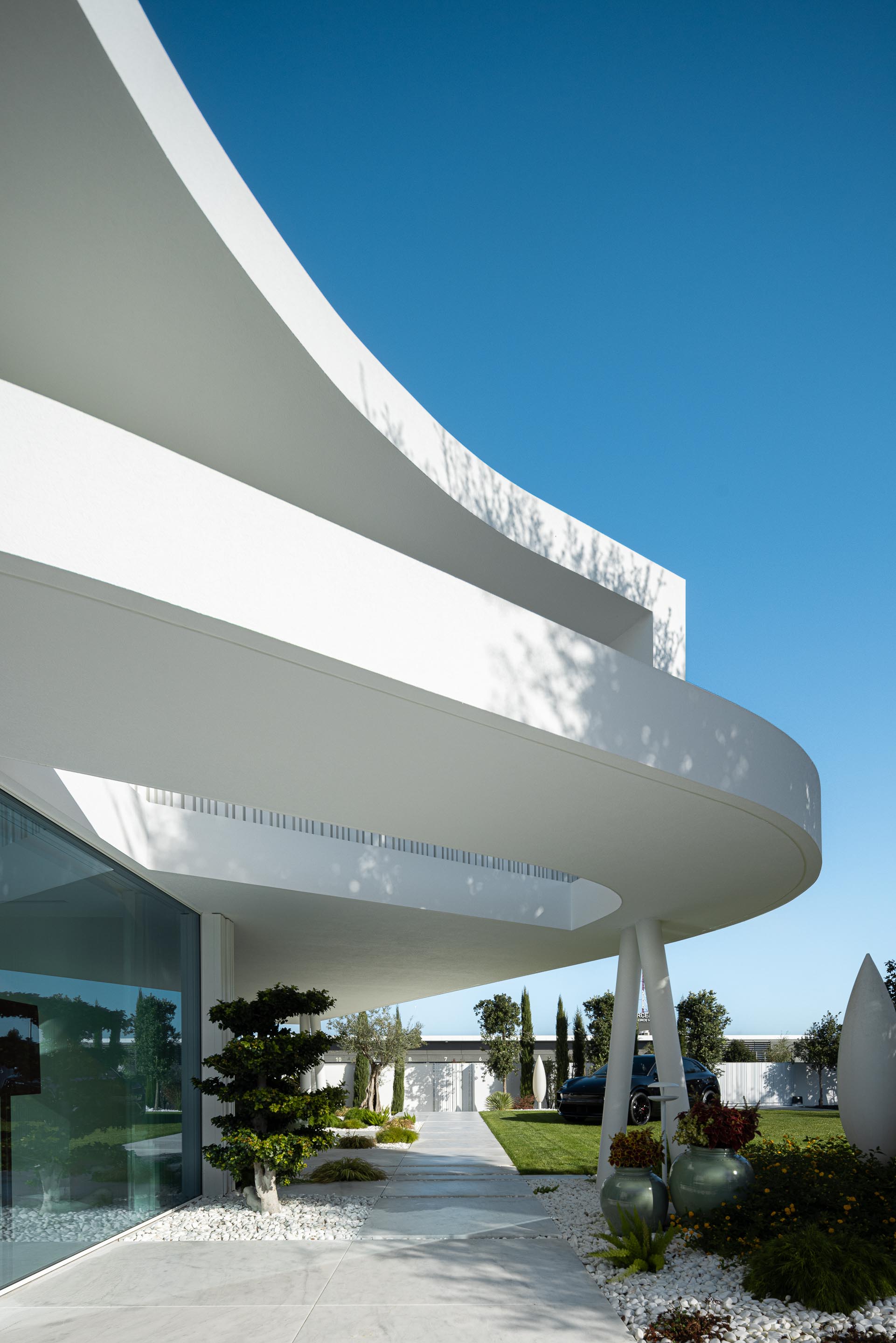 Moradia com varanda curva em consola e pilares esguios, ladeada por um percurso em pavimento claro e jardim — RiscoWhite arquitectura Risco Singular – Arquitectura, Lda. fotografia Ivo Tavares
