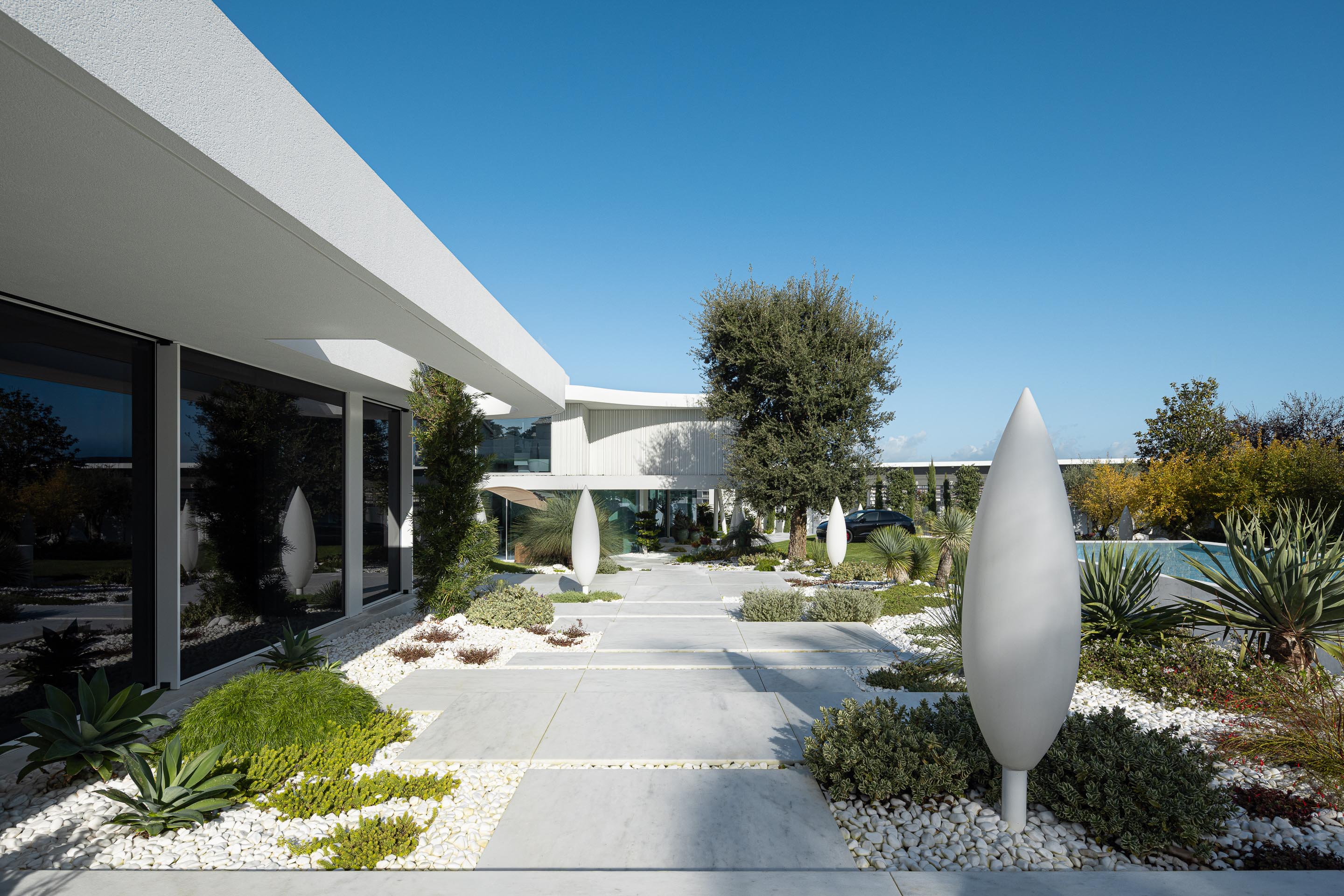 Percurso exterior em lajes de pedra clara, ladeado por jardim com seixos brancos, vegetação diversa e esculturas — RiscoWhite arquitectura Risco Singular – Arquitectura, Lda. fotografia Ivo Tavares