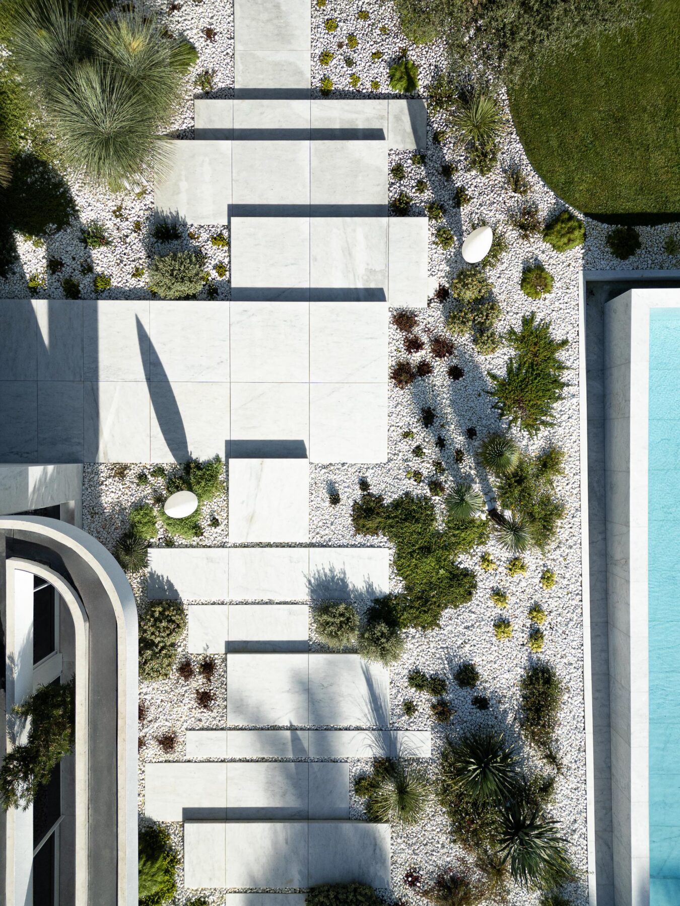 Vista aérea do jardim com pavimento de lajes claras, seixos brancos, vegetação densa e piscina de água azul — RiscoWhite arquitectura Risco Singular – Arquitectura, Lda. fotografia Ivo Tavares