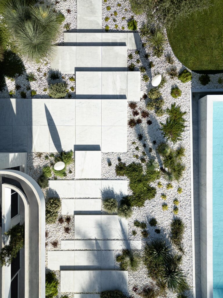 Vista aérea do jardim com pavimento de lajes claras, seixos brancos, vegetação densa e piscina de água azul — RiscoWhite arquitectura Risco Singular – Arquitectura, Lda. fotografia Ivo Tavares