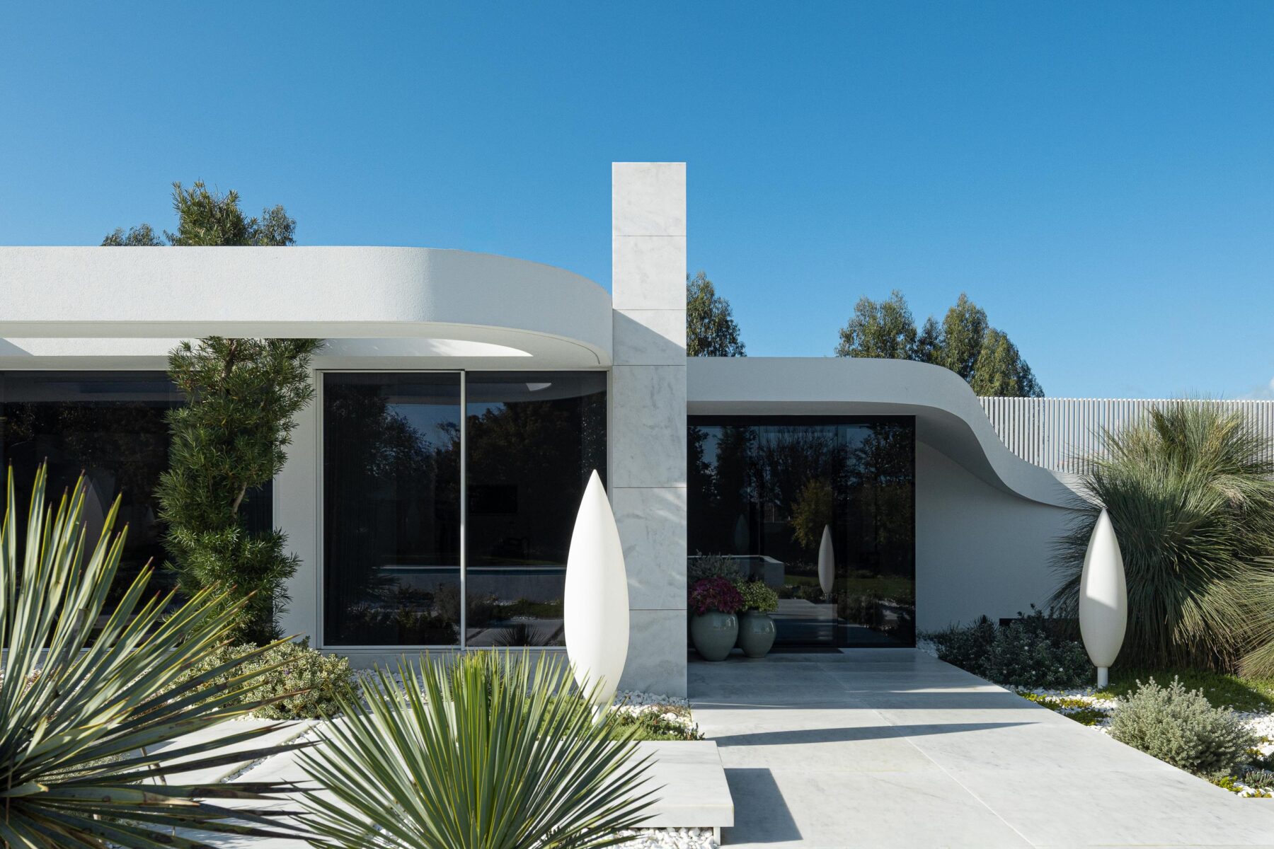 Entrada da moradia com pilar vertical em mármore branco e volumes curvos, enquadrada por vegetação exuberante — RiscoWhite arquitectura Risco Singular – Arquitectura, Lda. fotografia Ivo Tavares