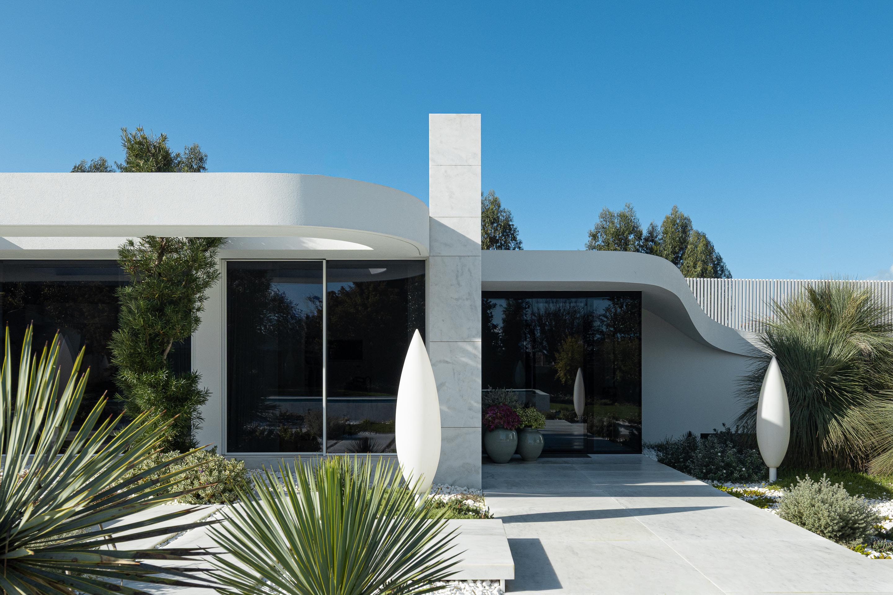 Entrada da moradia com pilar vertical em mármore branco e volumes curvos, enquadrada por vegetação exuberante — RiscoWhite arquitectura Risco Singular – Arquitectura, Lda. fotografia Ivo Tavares