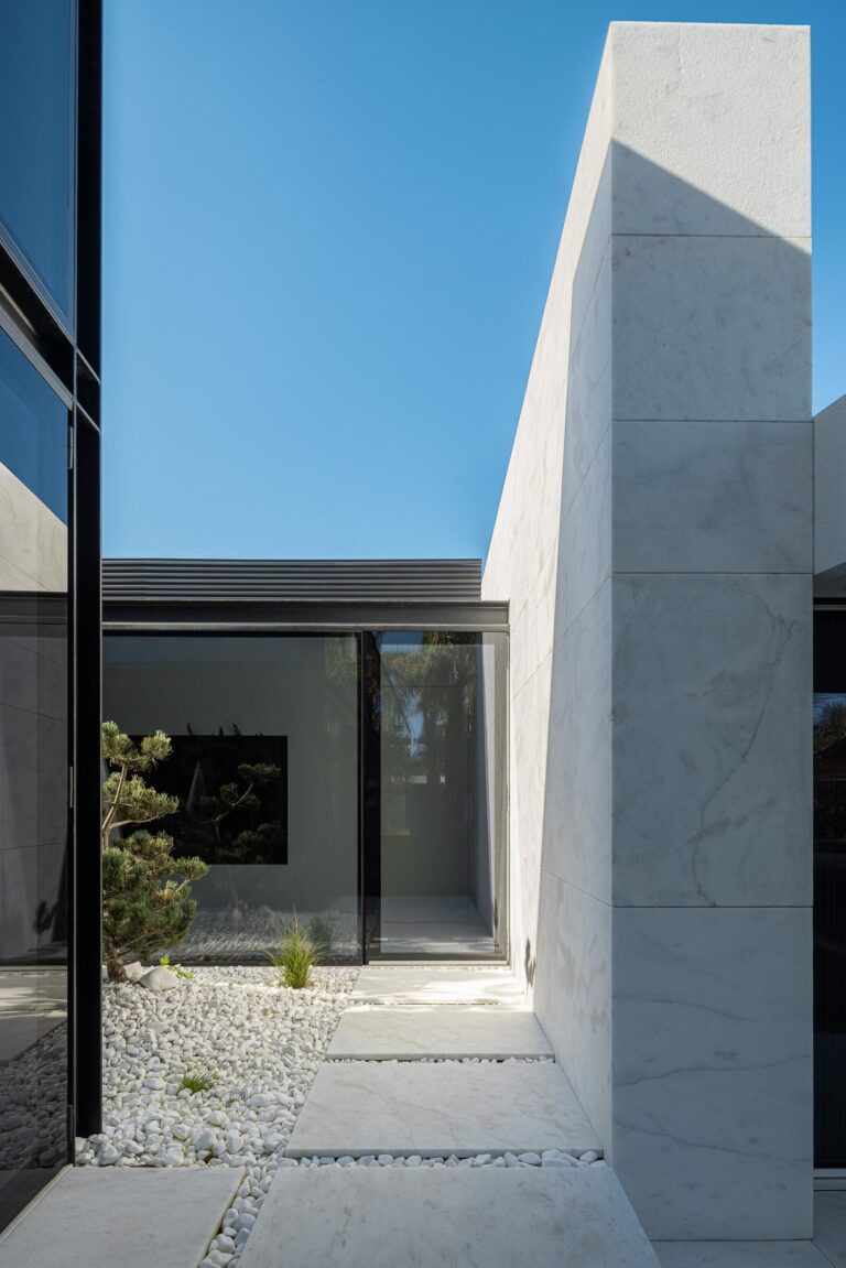 Corredor exterior entre volumes, com pilar em mármore branco, paredes envidraçadas e pavimento de seixos — RiscoWhite arquitectura Risco Singular – Arquitectura, Lda. fotografia Ivo Tavares